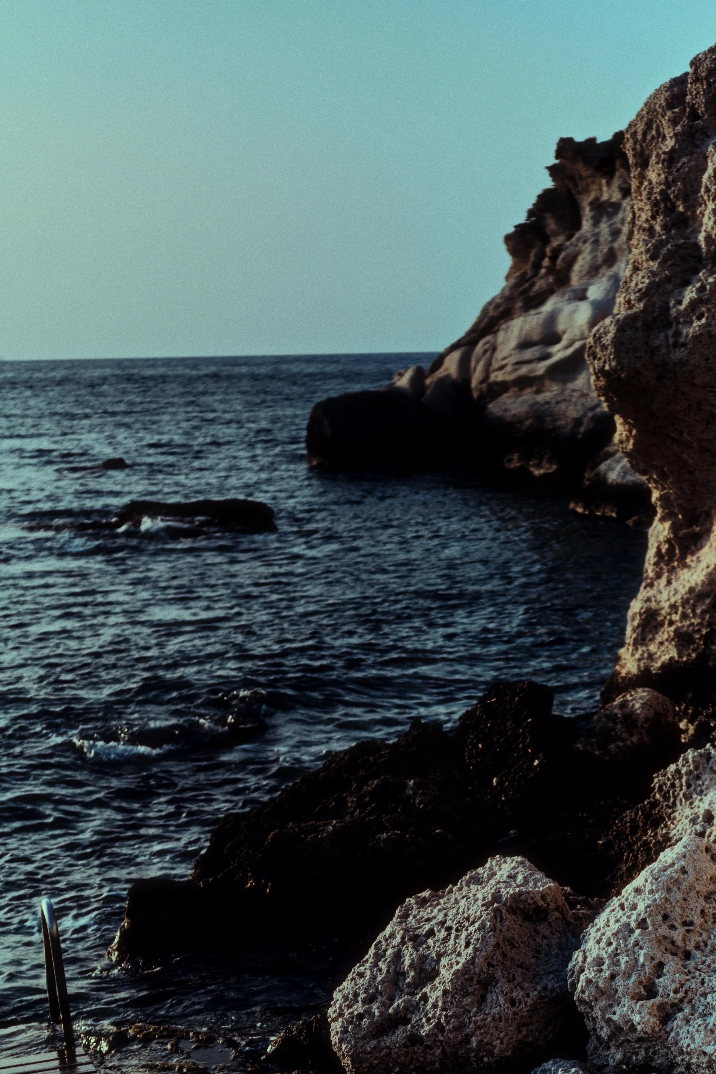 Rocky coastline at the beach with clear water and a pale sky in the background.