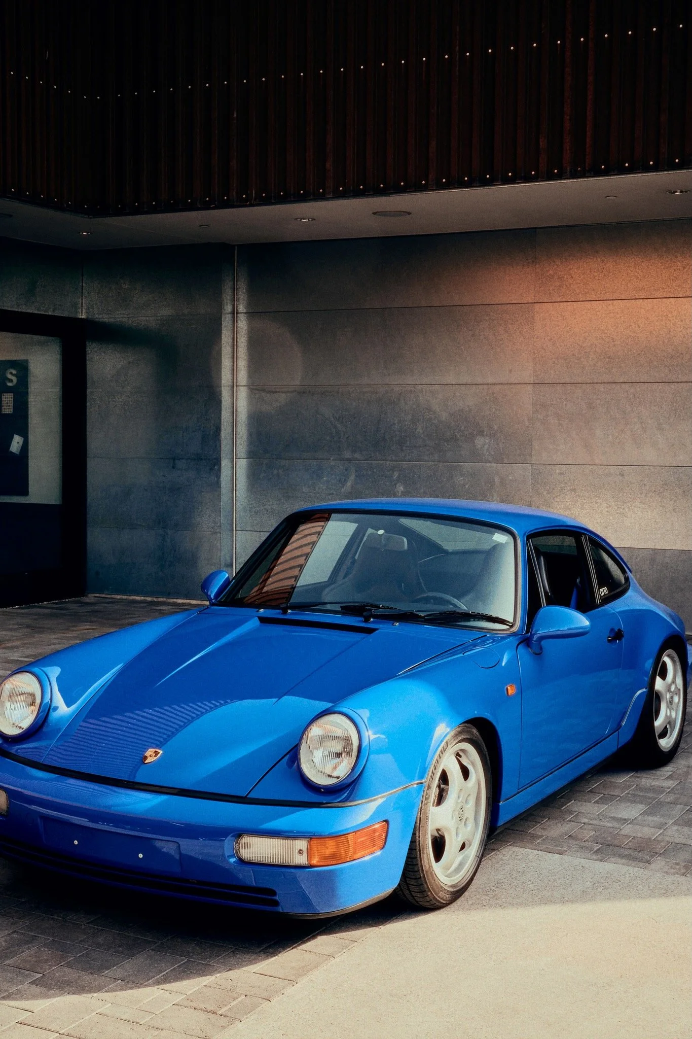 A blue vintage Porsche sports car parked outside a modern building with metallic walls.