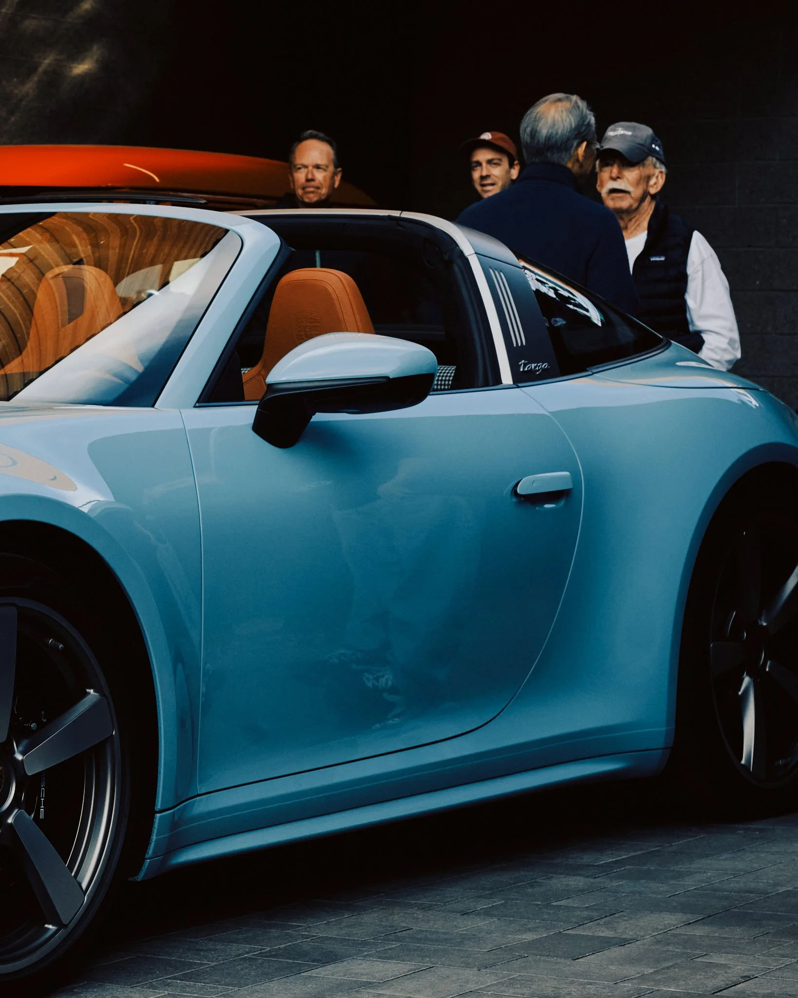 Light blue sports car with tan interior in an indoor setting, with four men standing behind it talking.