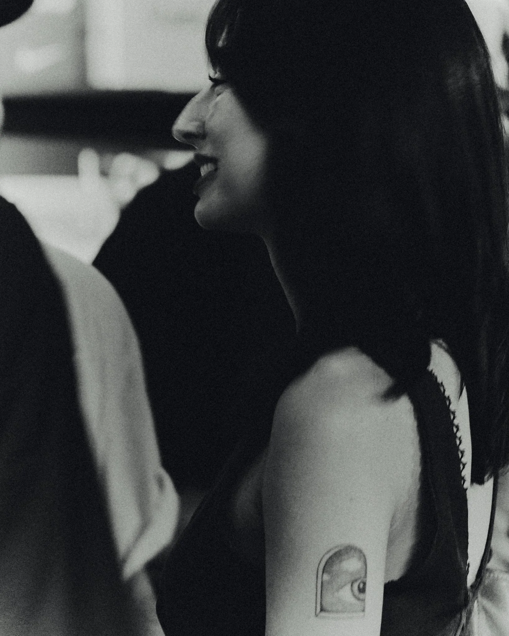 A woman smiling with dark hair, showing a shoulder tattoo of an eye inside an arch.