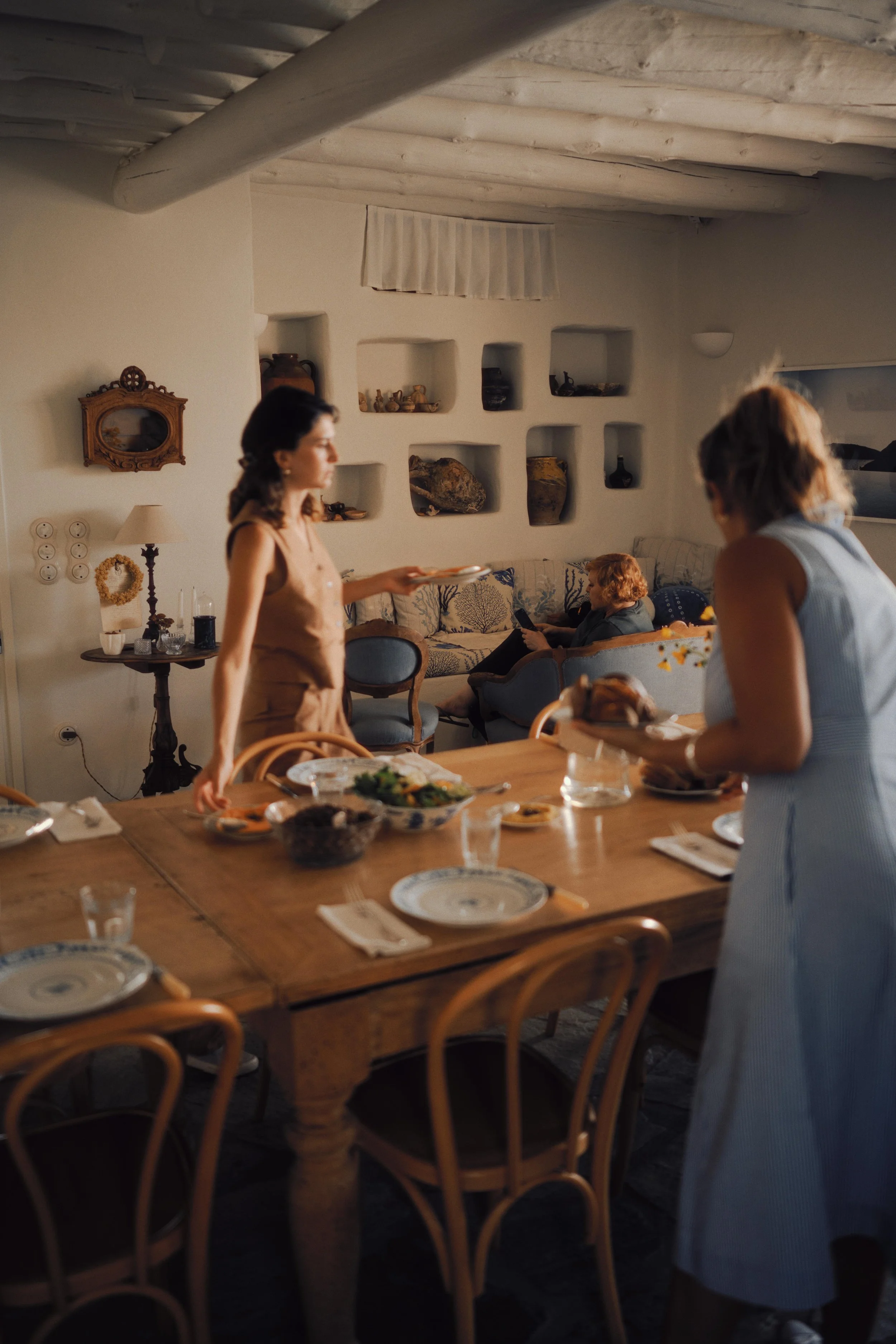 A family gathering in a cozy dining room with a wooden table set with plates, bowls, and glasses. Two women are serving food, and an elderly person is sitting on a couch in the background using a tablet. The room features built-in shelves with decora