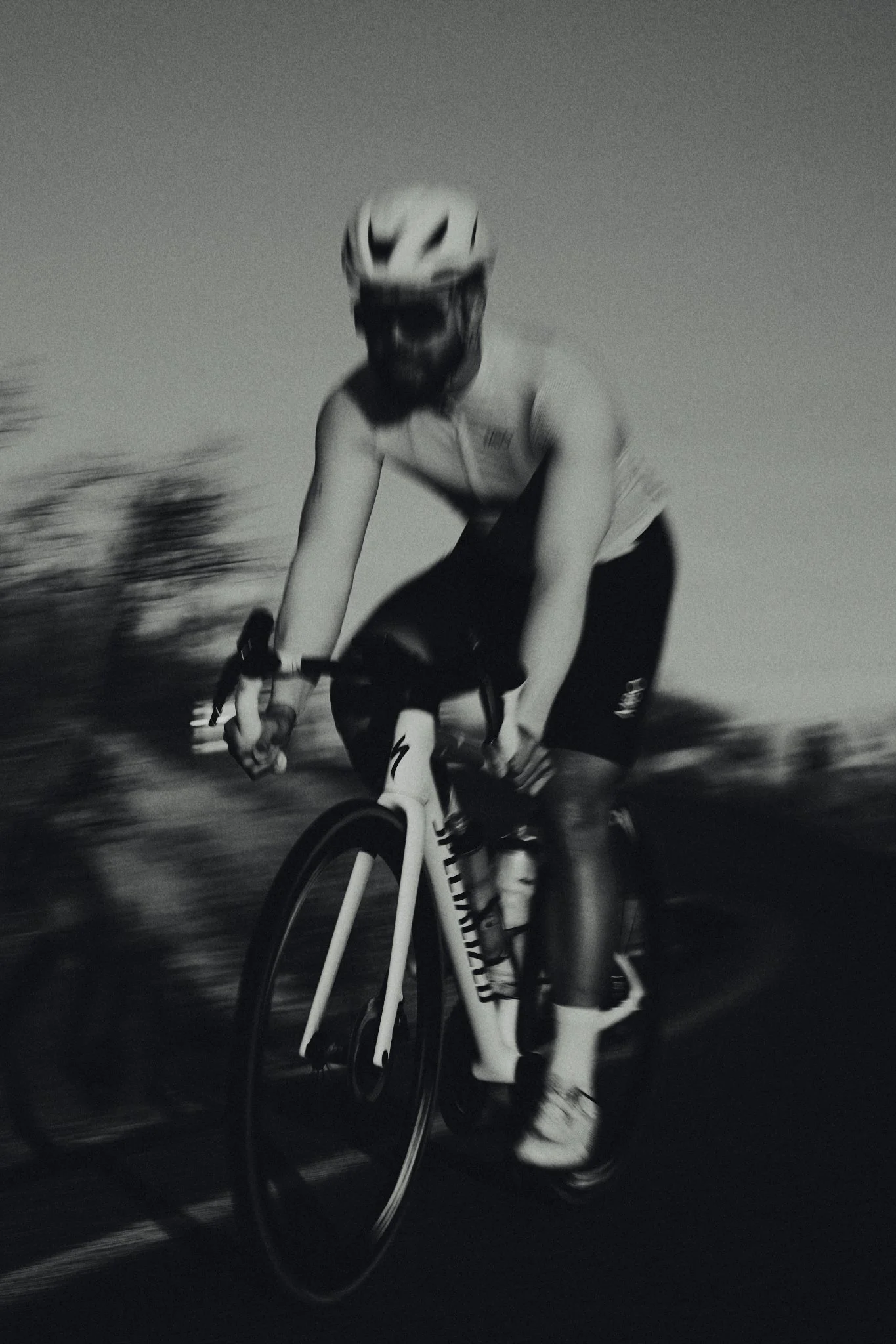 A person wearing a helmet and cycling gear rides a road bike outdoors during dusk or dawn in black and white.