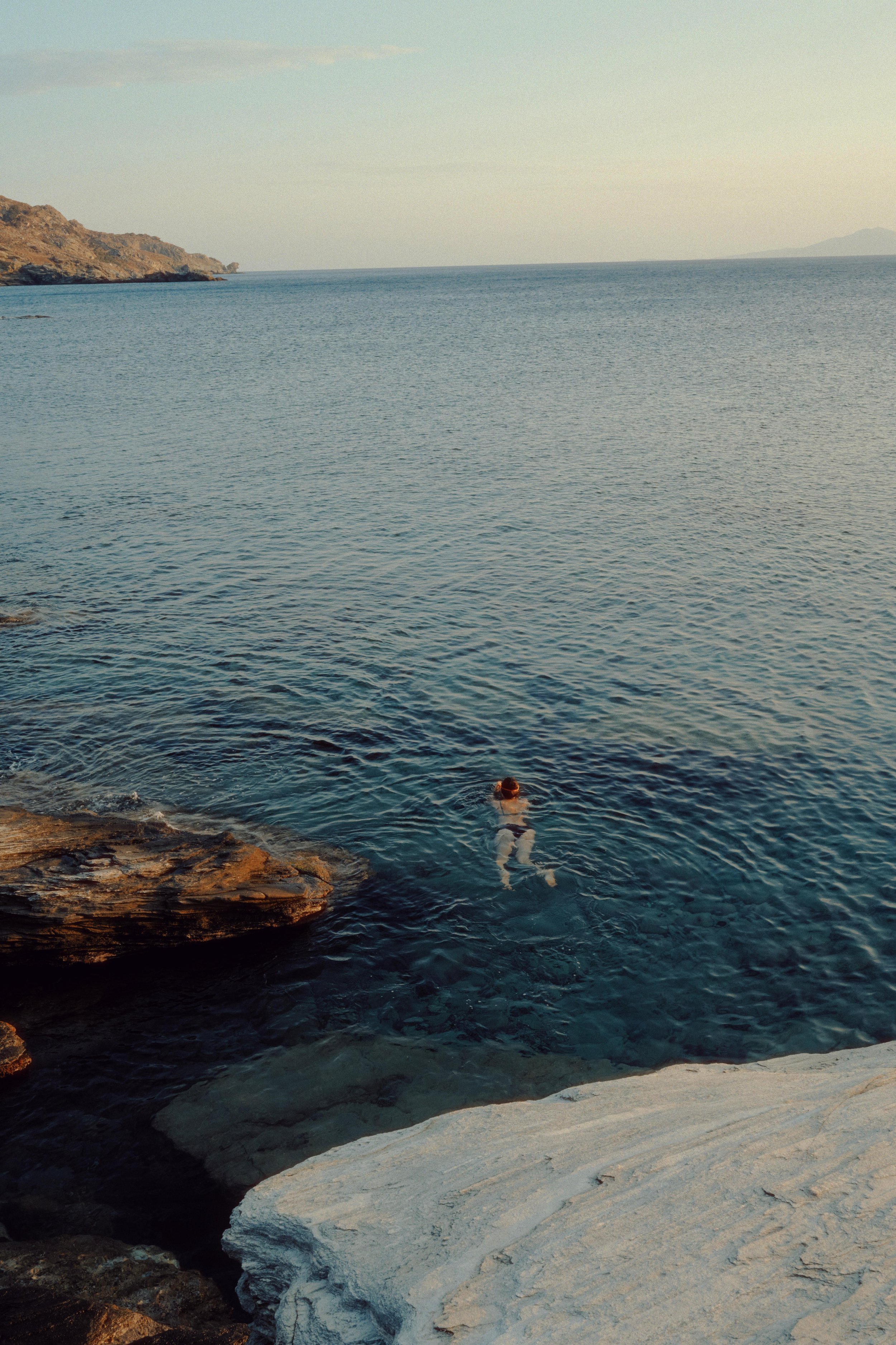 A person swimming in the ocean near rocky shores with a distant coastline and mountains in the background.