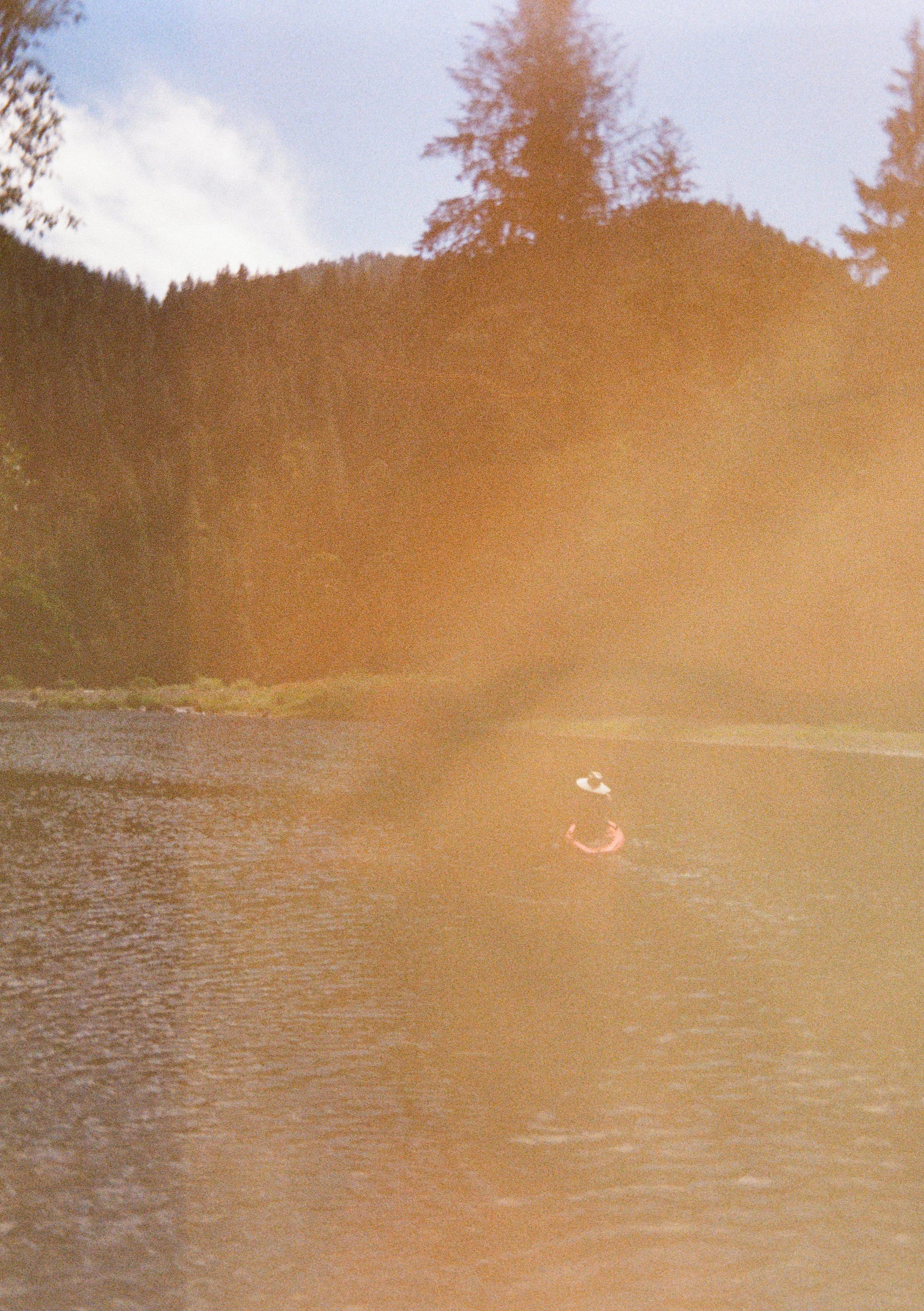 Person kayaking on a river surrounded by tall trees with a partly cloudy sky.