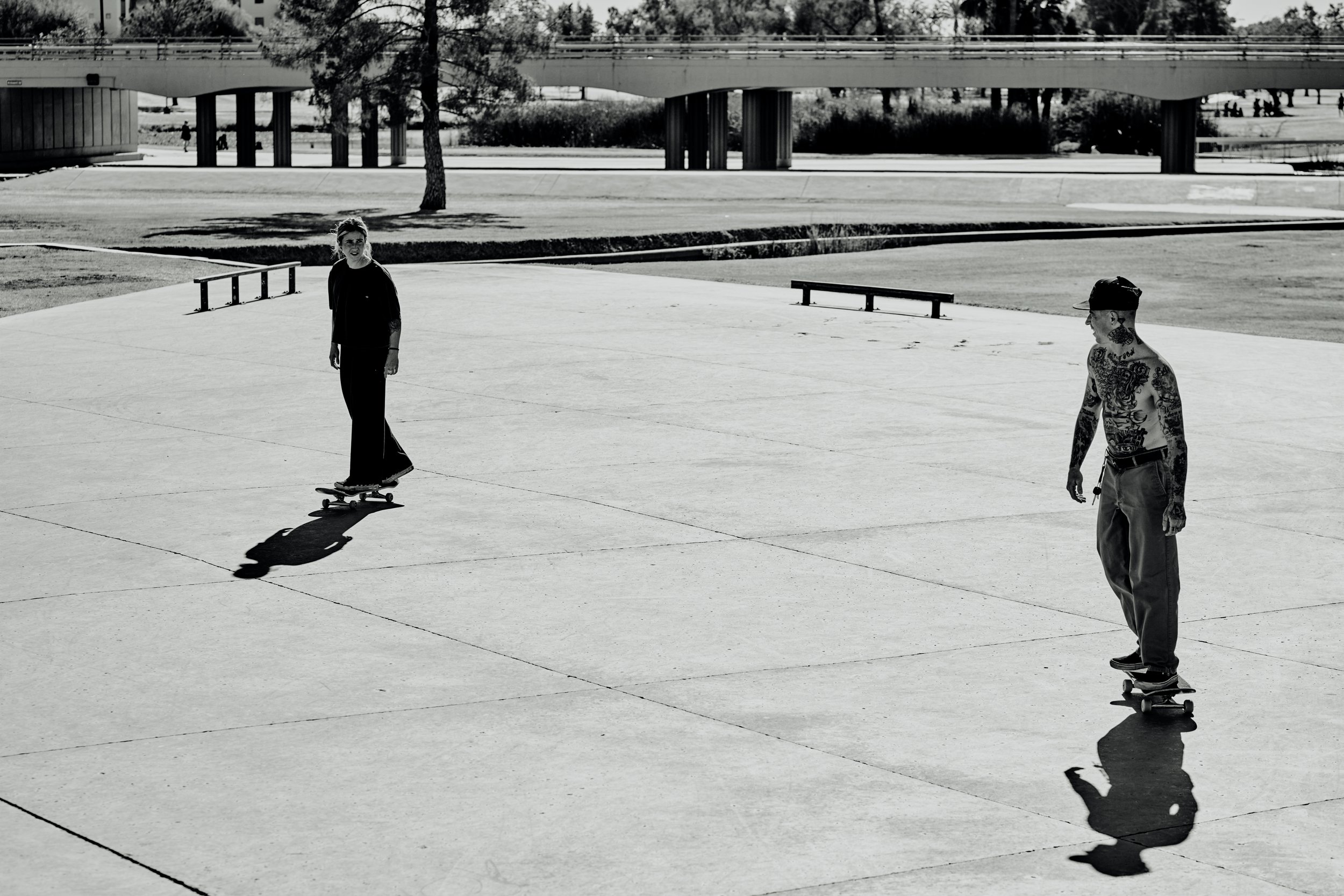 A woman skateboarding and a tattooed man skateboarding in an open concrete area with shadows cast on the ground and a bridge in the background.