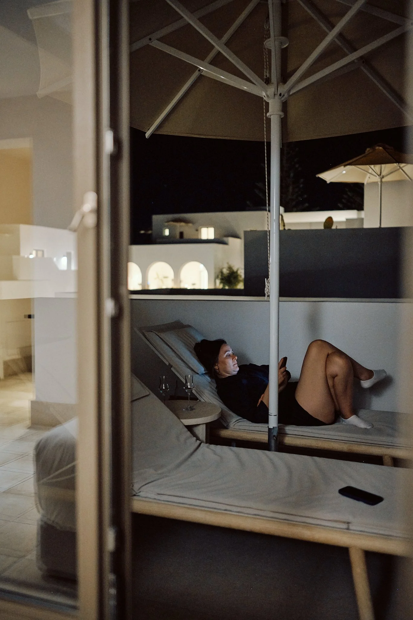 A woman lying on a lounge chair outside at night, looking at her phone, with a glass of wine nearby, under an umbrella on a balcony or terrace with white buildings in the background.