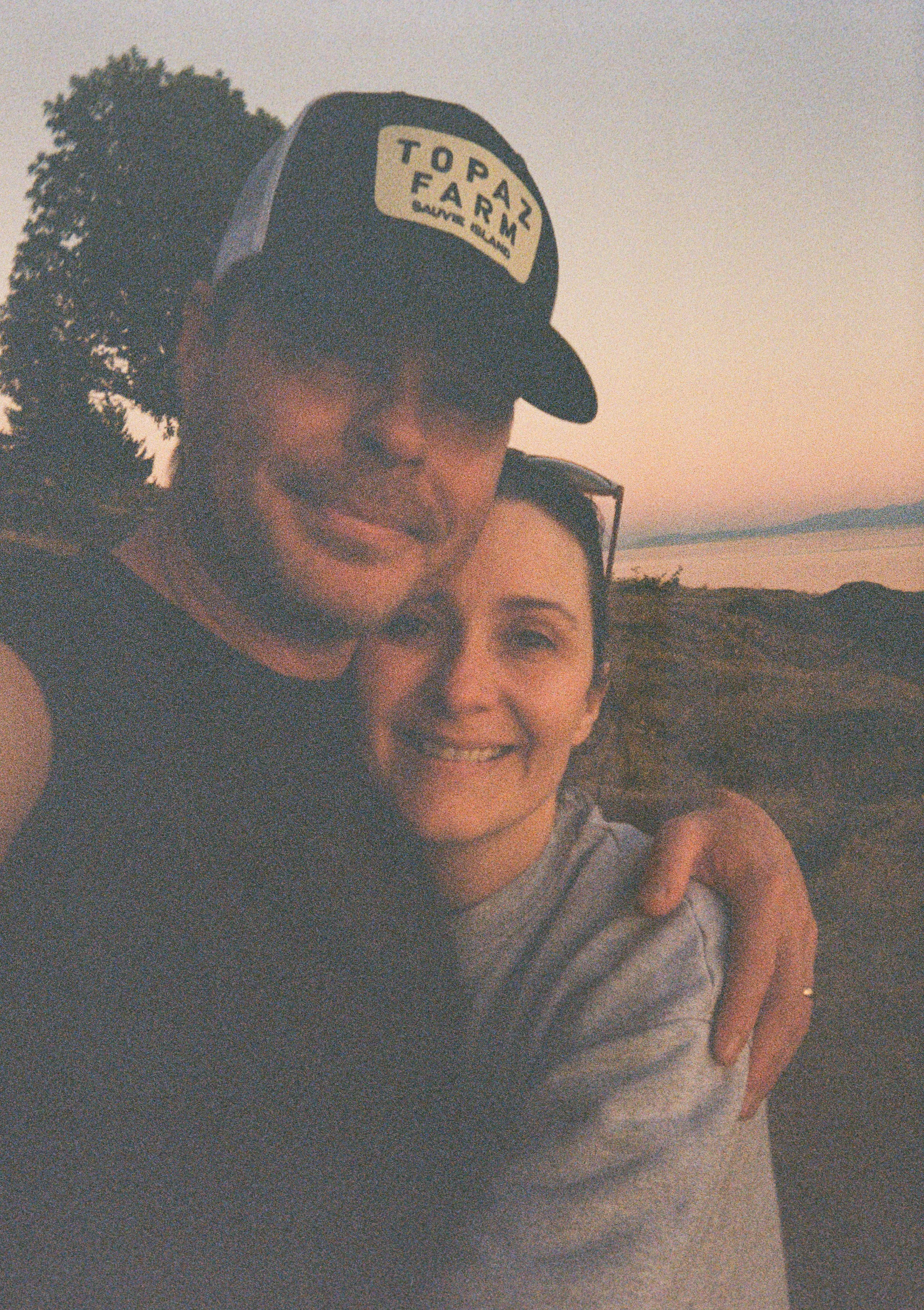 A man and woman smiling and hugging outdoors during sunset, with a body of water and a tree in the background.