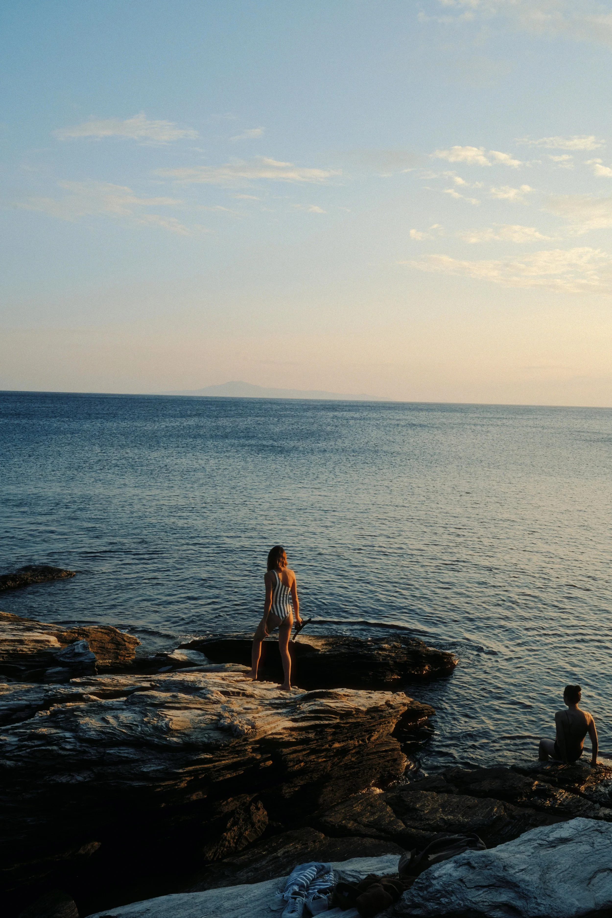 Two people sitting on rocks by the ocean during sunset, with one person standing and holding an object, and the other sitting on a rock facing the water.