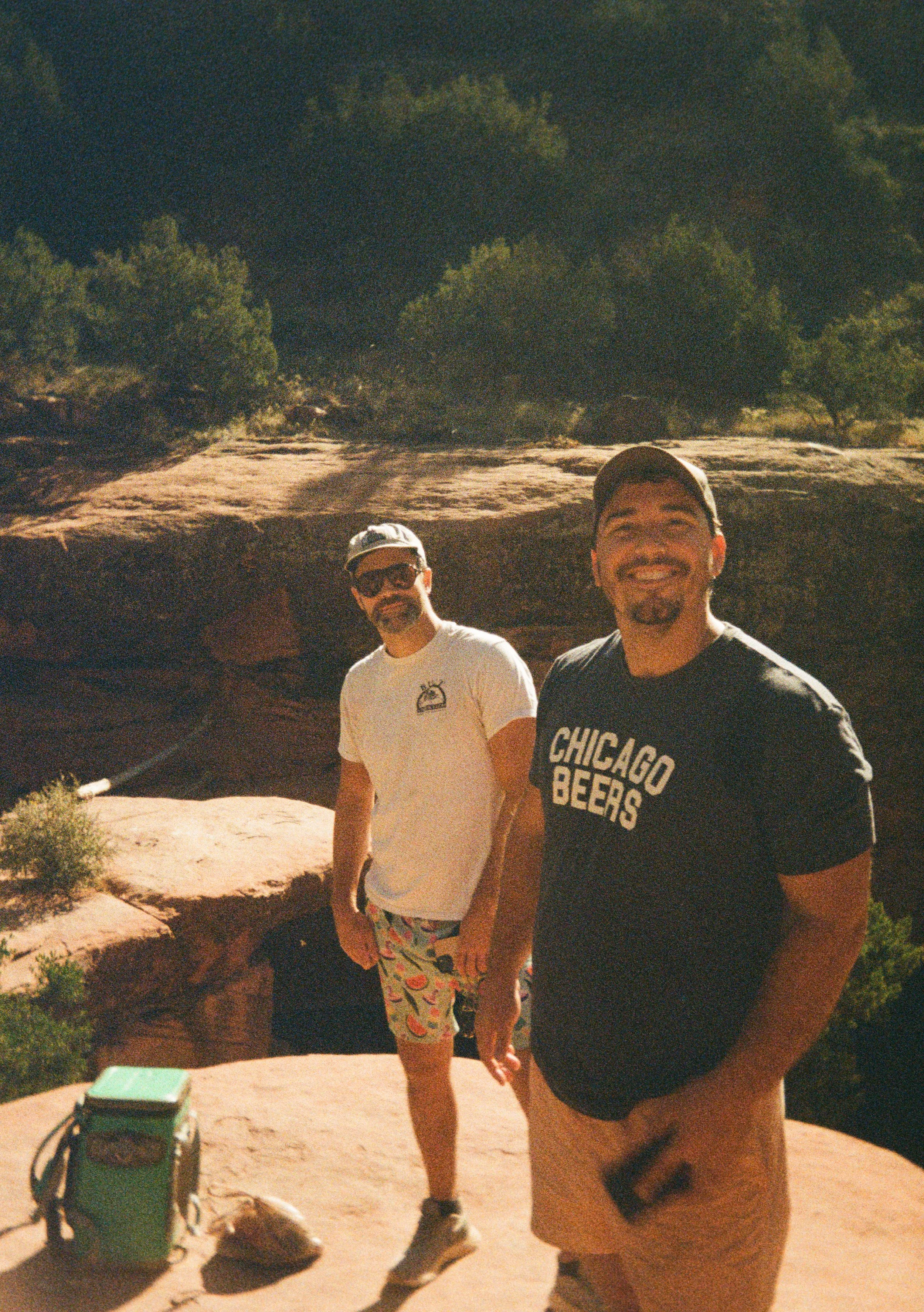 Two smiling men standing outdoors on a rocky ledge with a desert landscape and trees in the background during daytime.