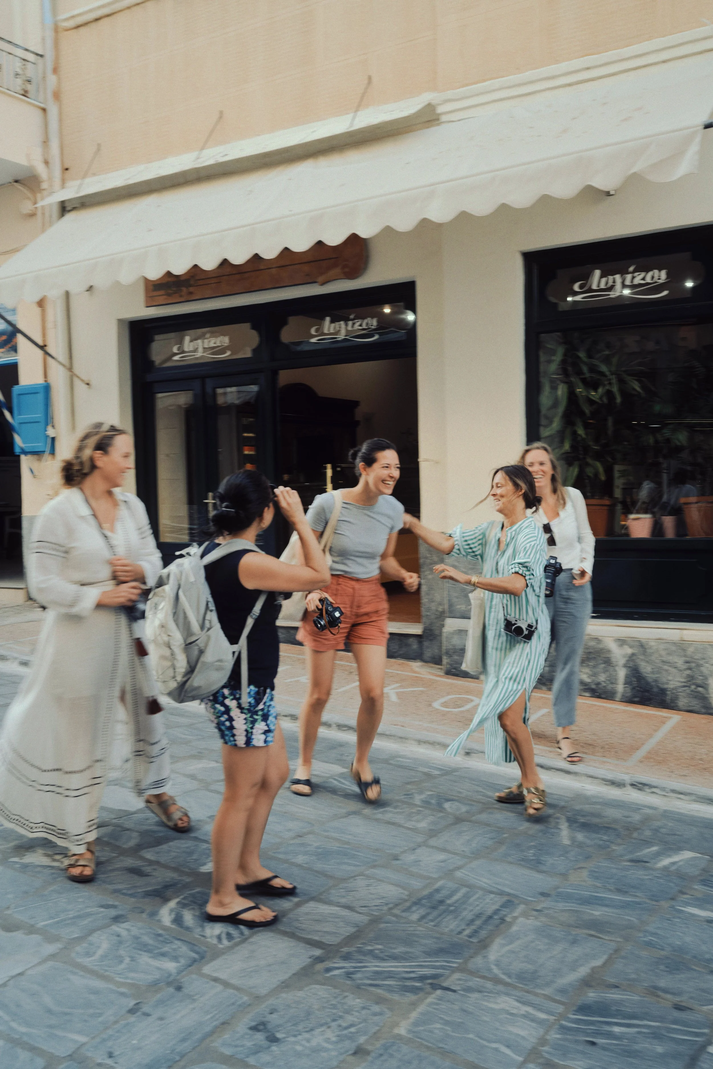 A group of six women laughing and chatting on a cobblestone street outside a café or restaurant, some holding cameras, dressed in casual summer clothing.
