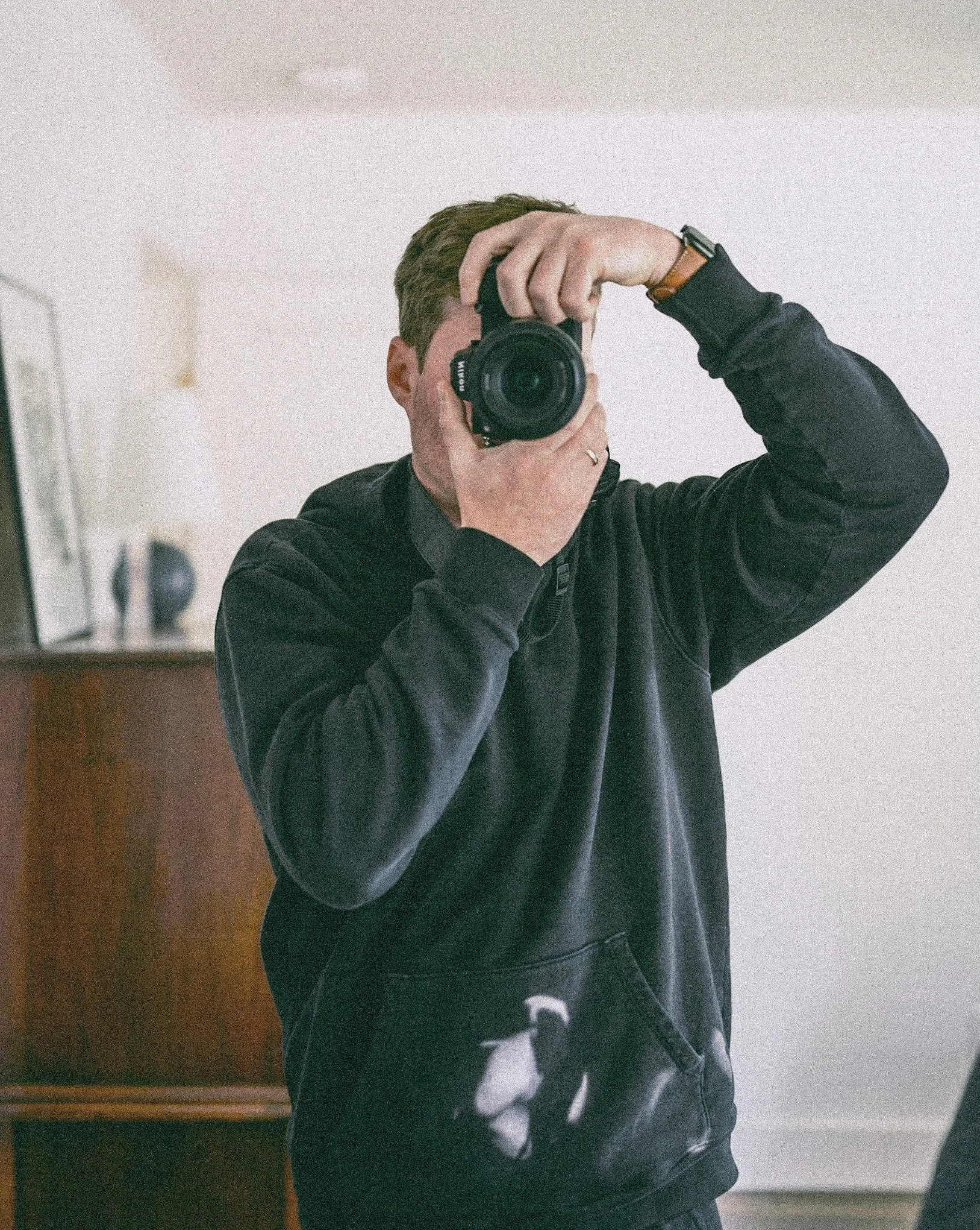 Person taking a mirror selfie with a camera, dressed in a black hoodie, with a white design on the pants, indoors with a mirror and furniture in the background.