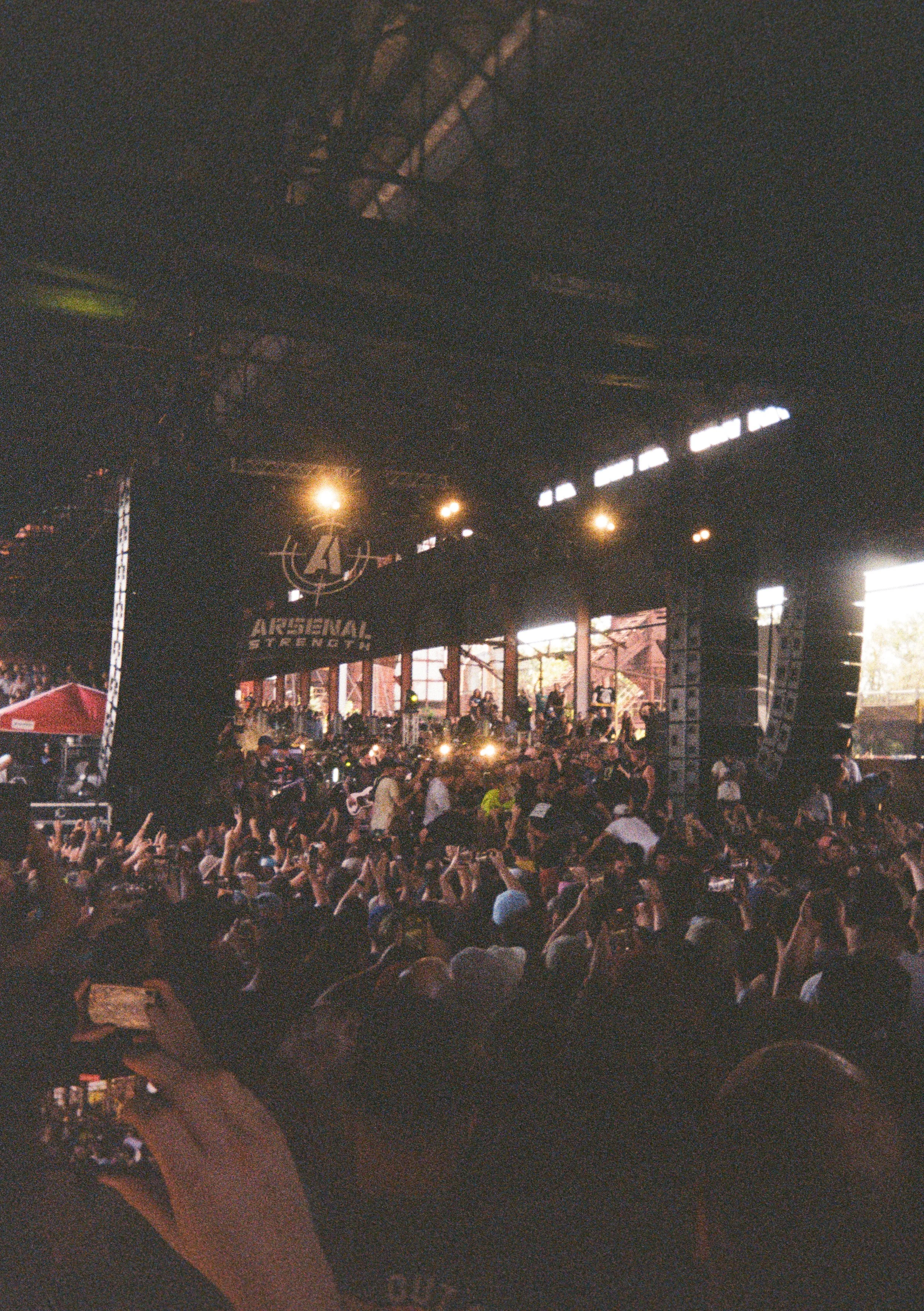 A crowded indoor concert with a stage labeled 'Arsenal Strength' and a large audience, many raising their hands, during a performance or event.