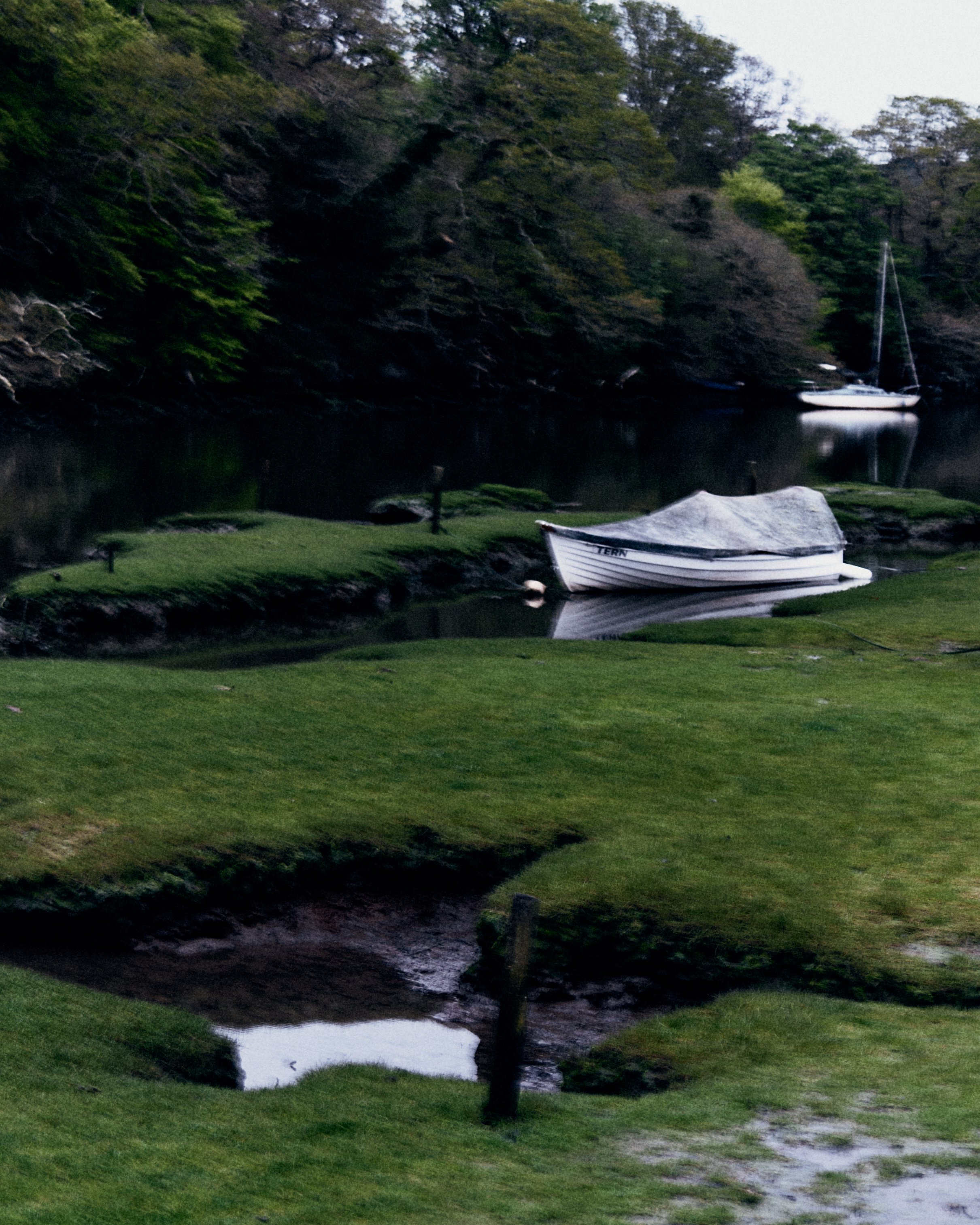 Two boats, one covered with a tarp, resting on a small waterway with lush green grass and trees in the background.