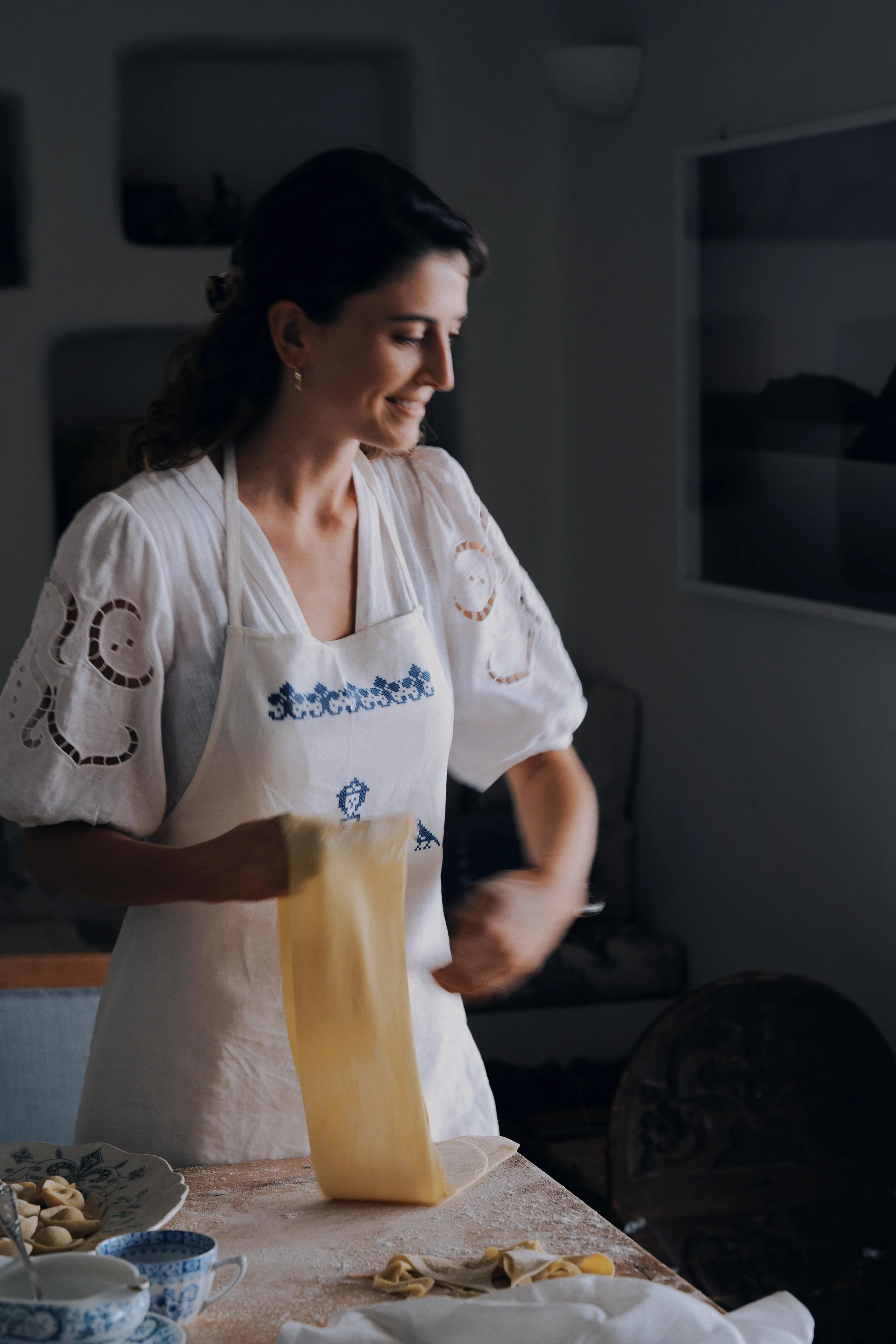 A woman in a white embroidered blouse and apron is in a kitchen, lifting a sheet of pasta dough, smiling as she prepares homemade pasta.