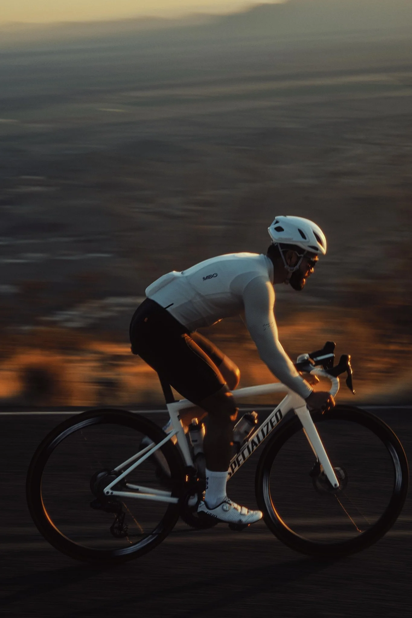 A person riding a road bicycle wearing a helmet and athletic gear during sunset, with a blurred background of the ocean or a large body of water.