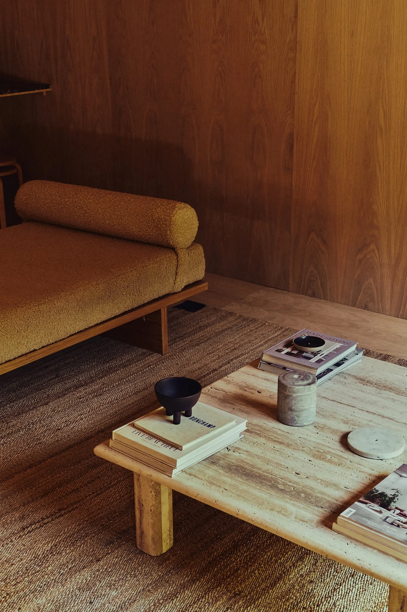 A cozy living room corner with a wooden wall, a yellow textured ottoman sofa, a low stone coffee table with stacked magazines, small black bowls, and decorative objects, on a multicolored woven rug.