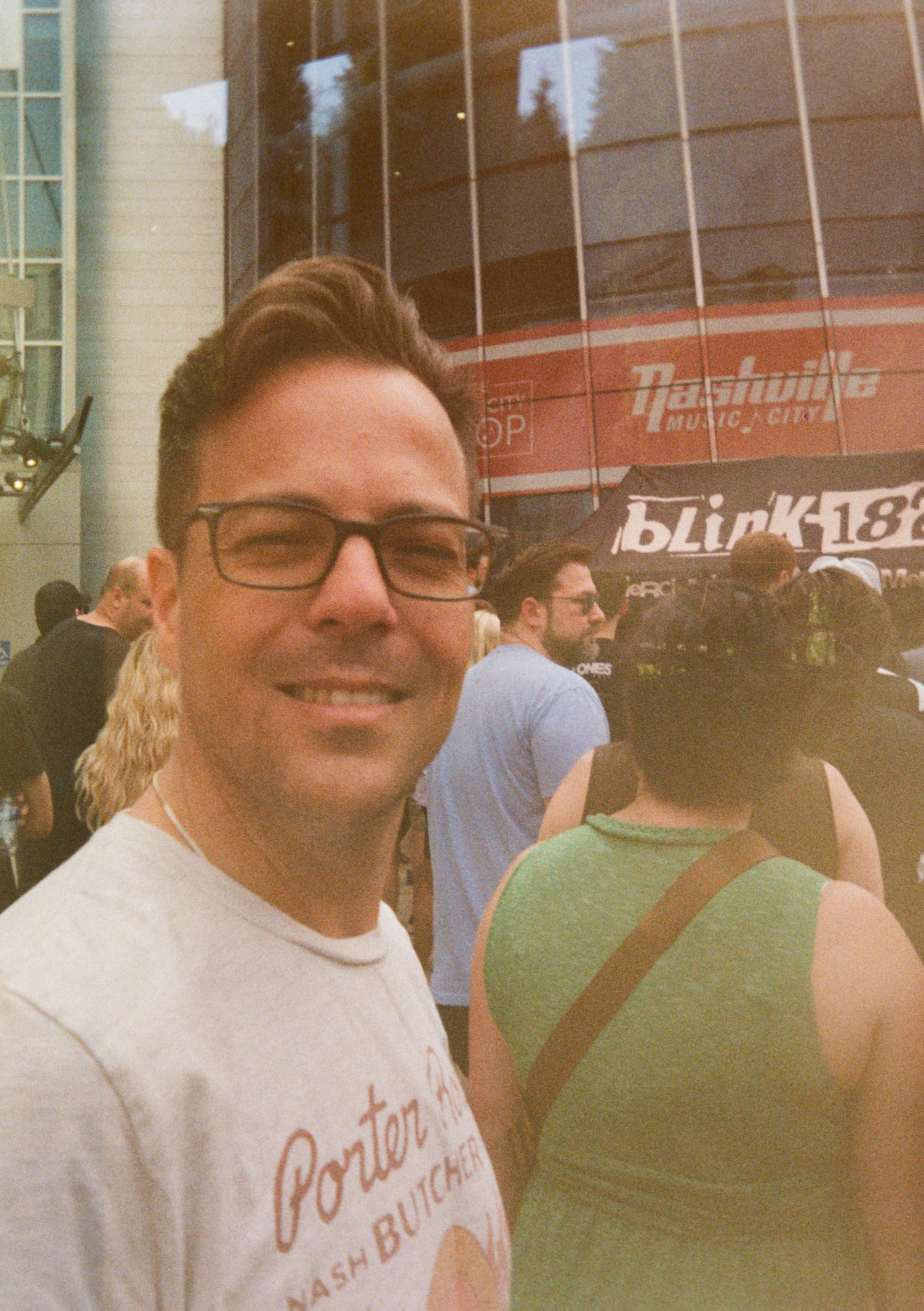 A man with glasses and a white T-shirt smiling in a crowded outdoor area, with a backdrop of a glass building and a music event banner.