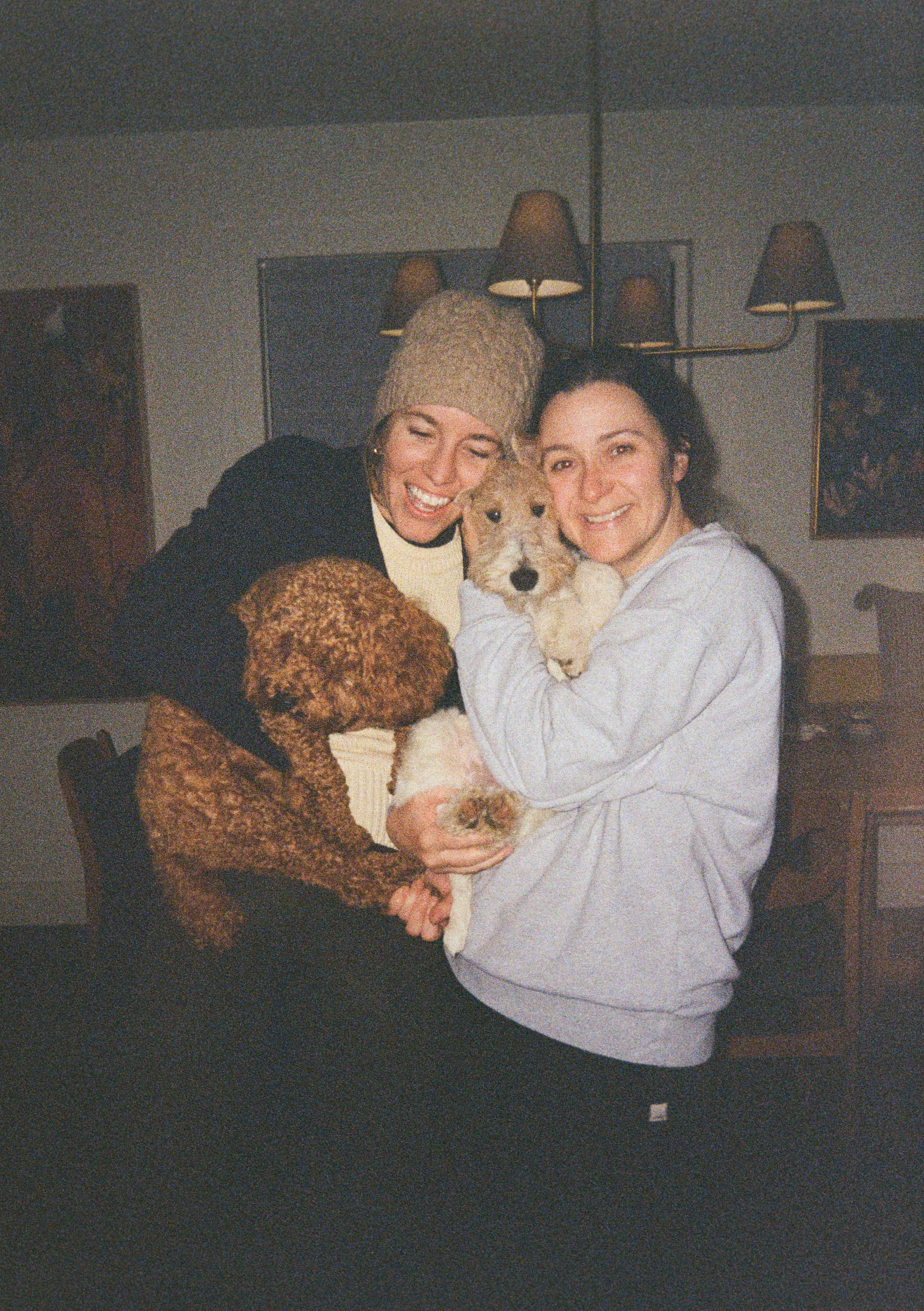 Two women smiling and hugging each other, holding three small dogs, in a cozy indoor setting.
