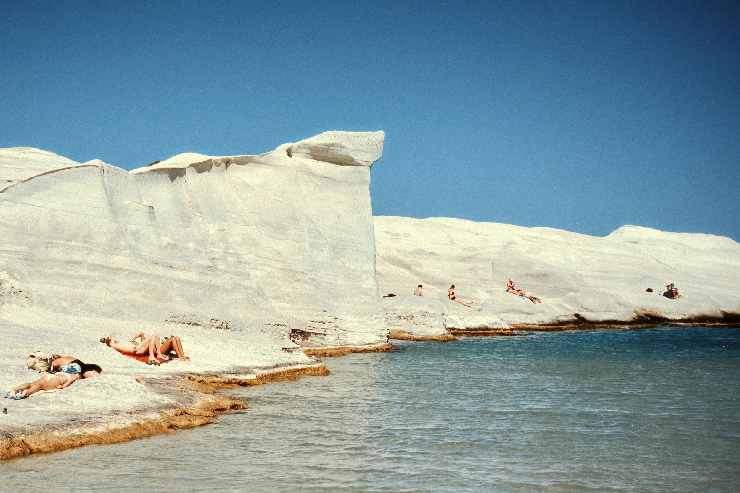People lounging and sunbathing on white rocky cliffs by the water under a clear blue sky.