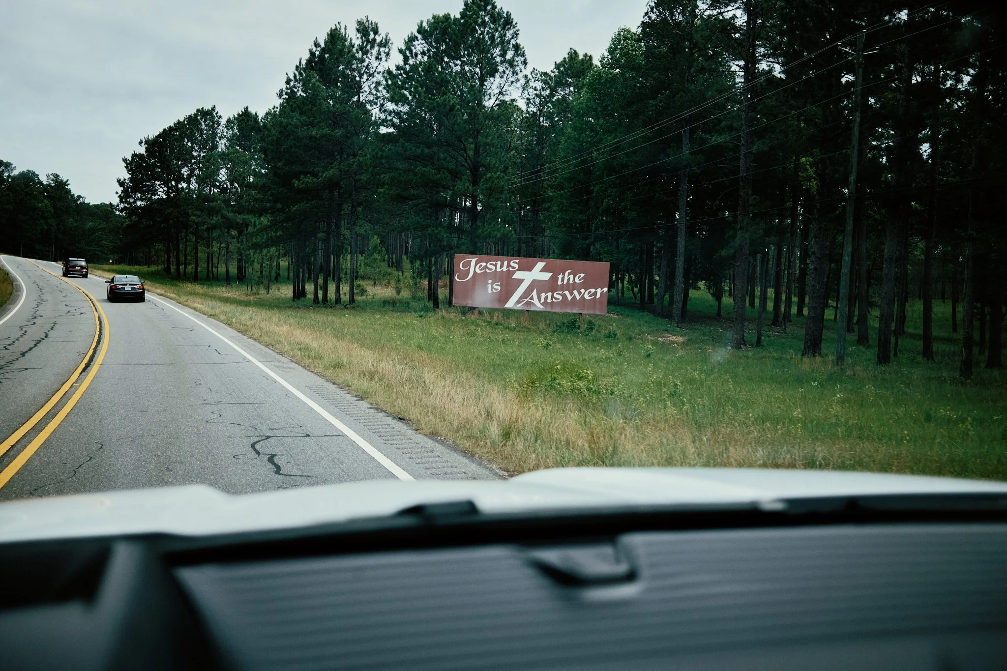View from inside a vehicle on a two-lane road with a Christian billboard on the side that reads 'Jesus is the Answer' amidst trees and power lines.