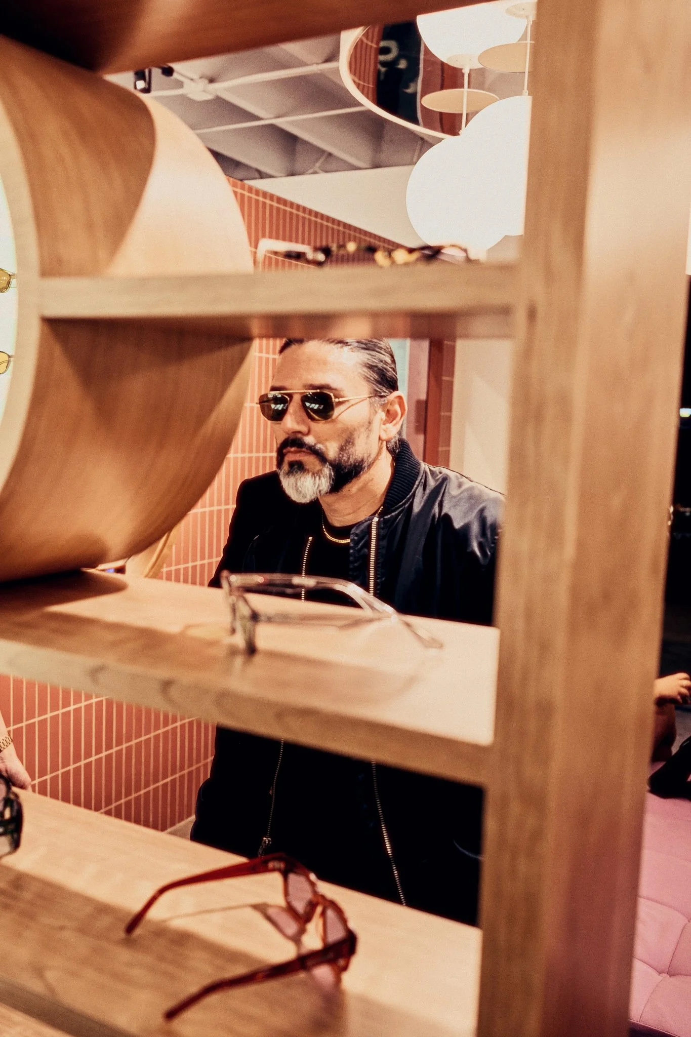 A man with a beard and sunglasses looking at eyeglasses in a store. The image is taken through a wooden display with glasses, with a pink tiled wall and modern lighting in the background.
