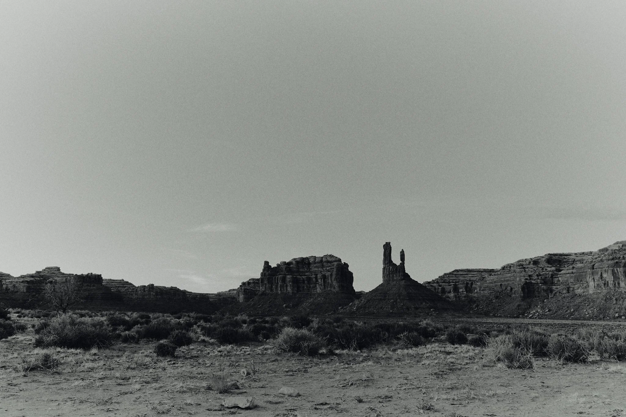 A black and white photo of a desert landscape with large rock formations and sparse desert vegetation, under a mostly clear sky.
