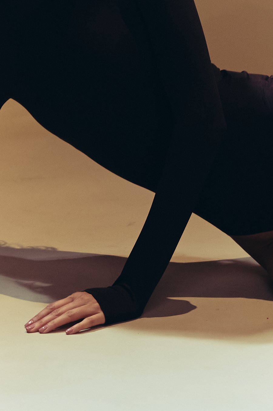 A person is on all fours on a beige surface, wearing black clothing, with one hand flat on the ground and the other bent, partially visible at the top right of the image.