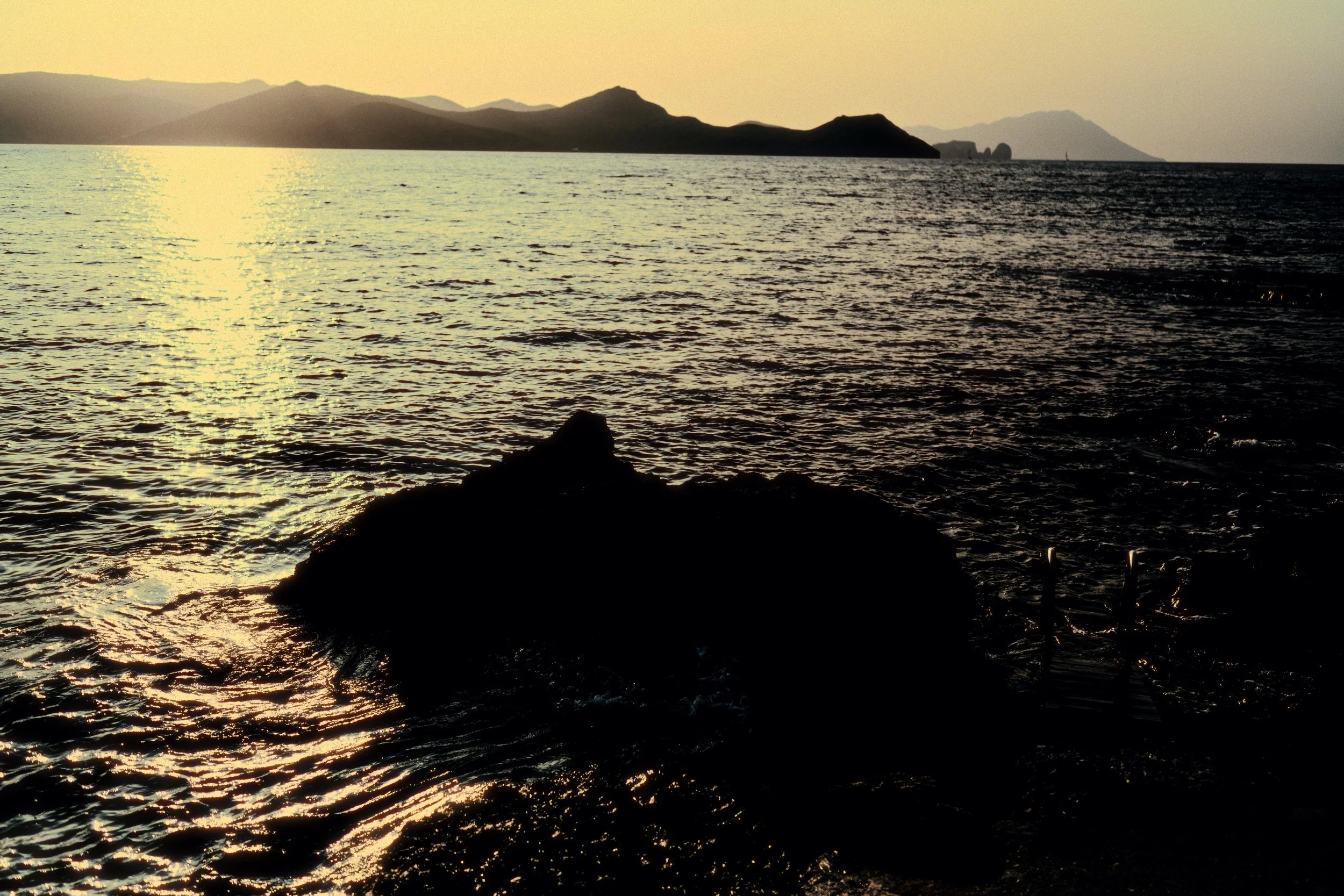 Sunset over ocean with mountainous landscape in the background, rocky shoreline in foreground, and calm water reflecting the sunlight.