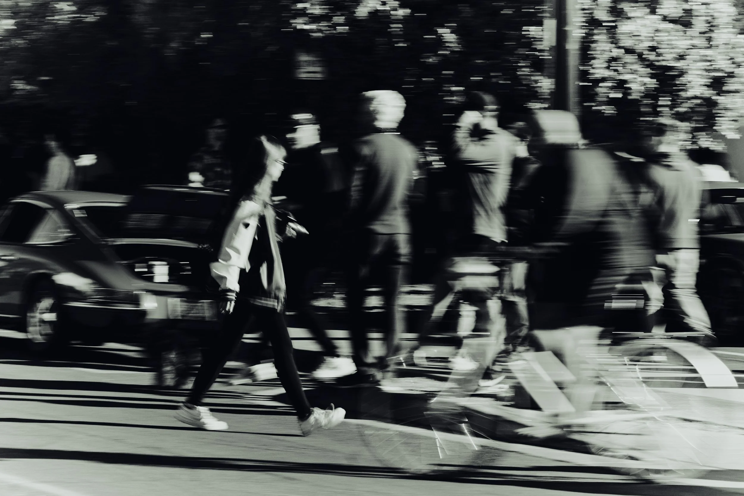 A black and white photo showing people crossing a street, with some walking and some standing, and cars parked along the side of the road, with blurred motion effects.