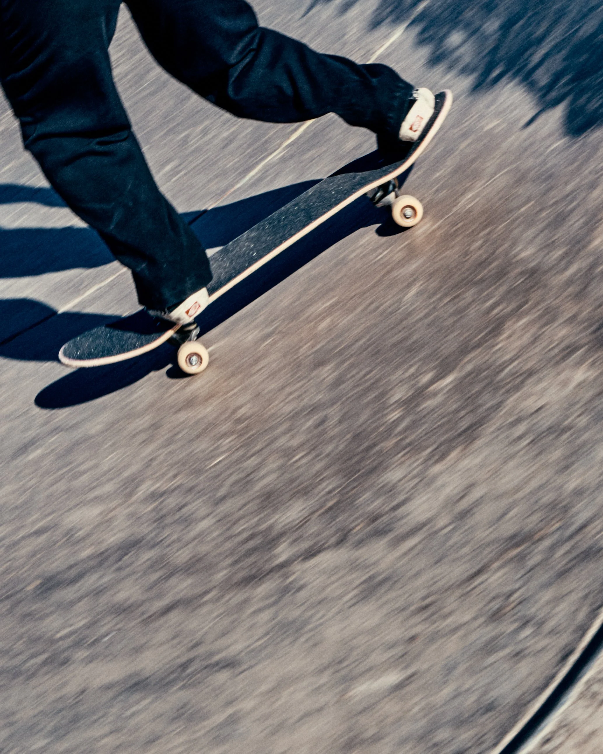 Close-up of a person riding a skateboard on a paved surface, wearing black pants and sneakers, with the skateboard angled as they push forward.