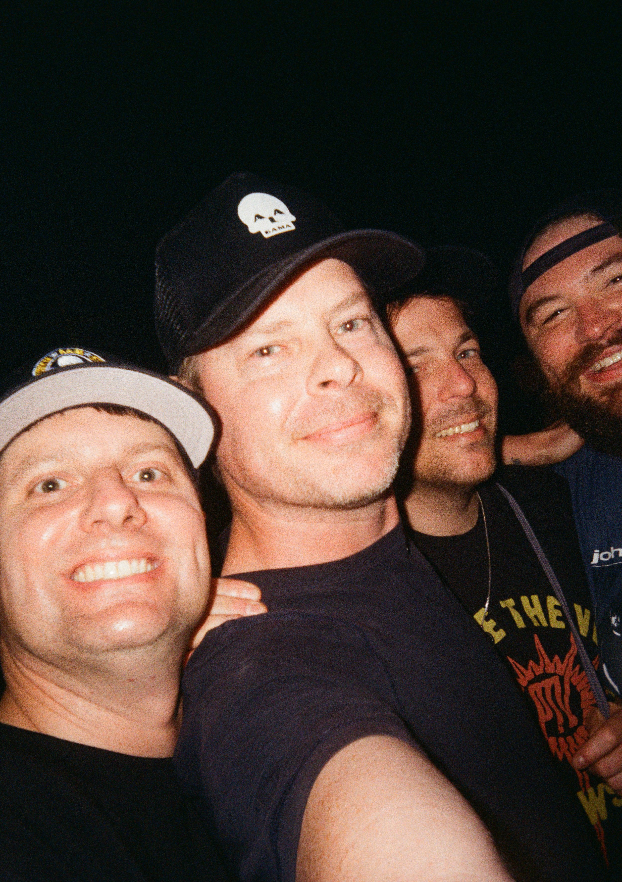 A group of four men taking a selfie together, smiling and wearing casual hats and shirts.