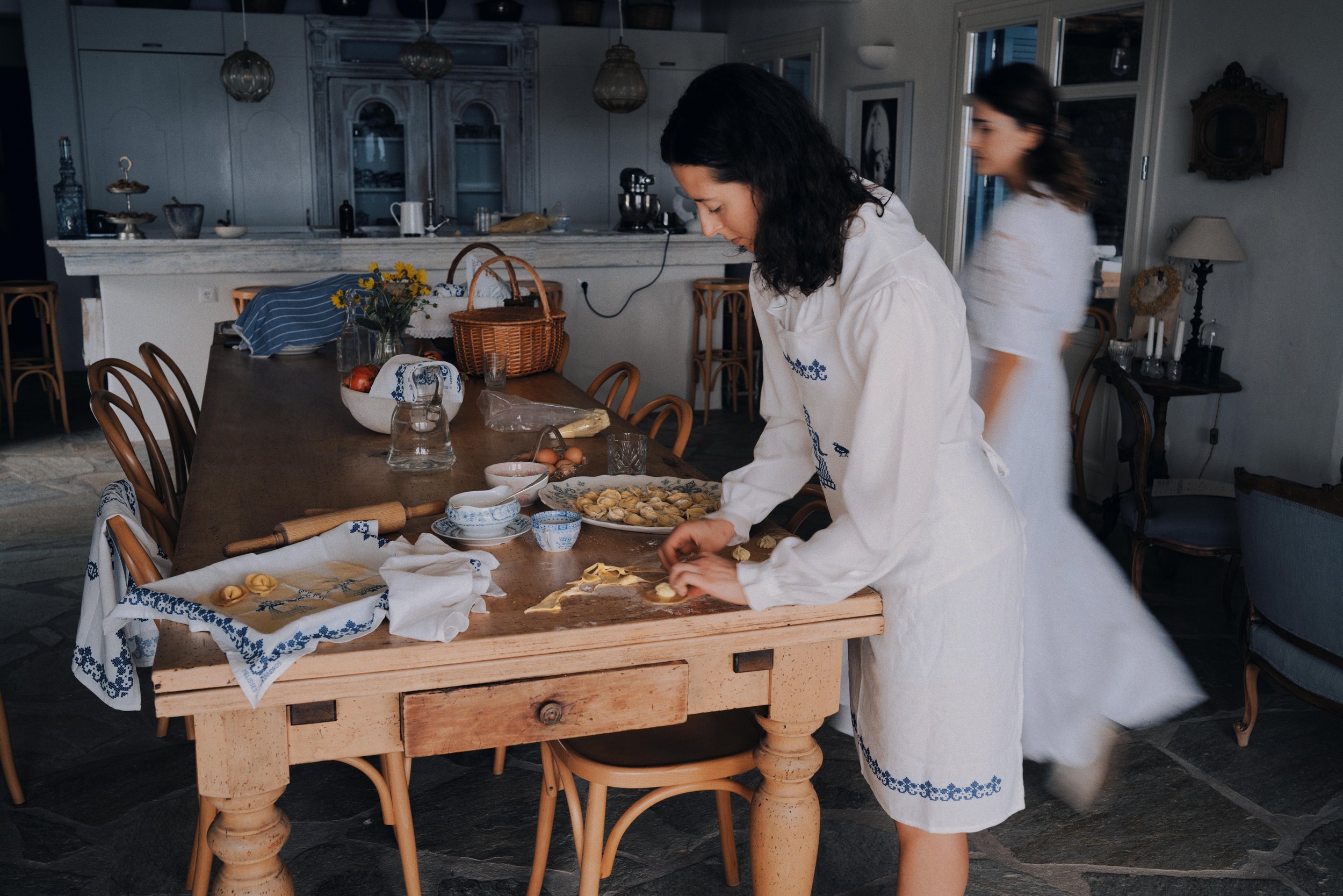 Two women preparing homemade pasta in a rustic kitchen, with various ingredients, utensils, and a large wooden table.