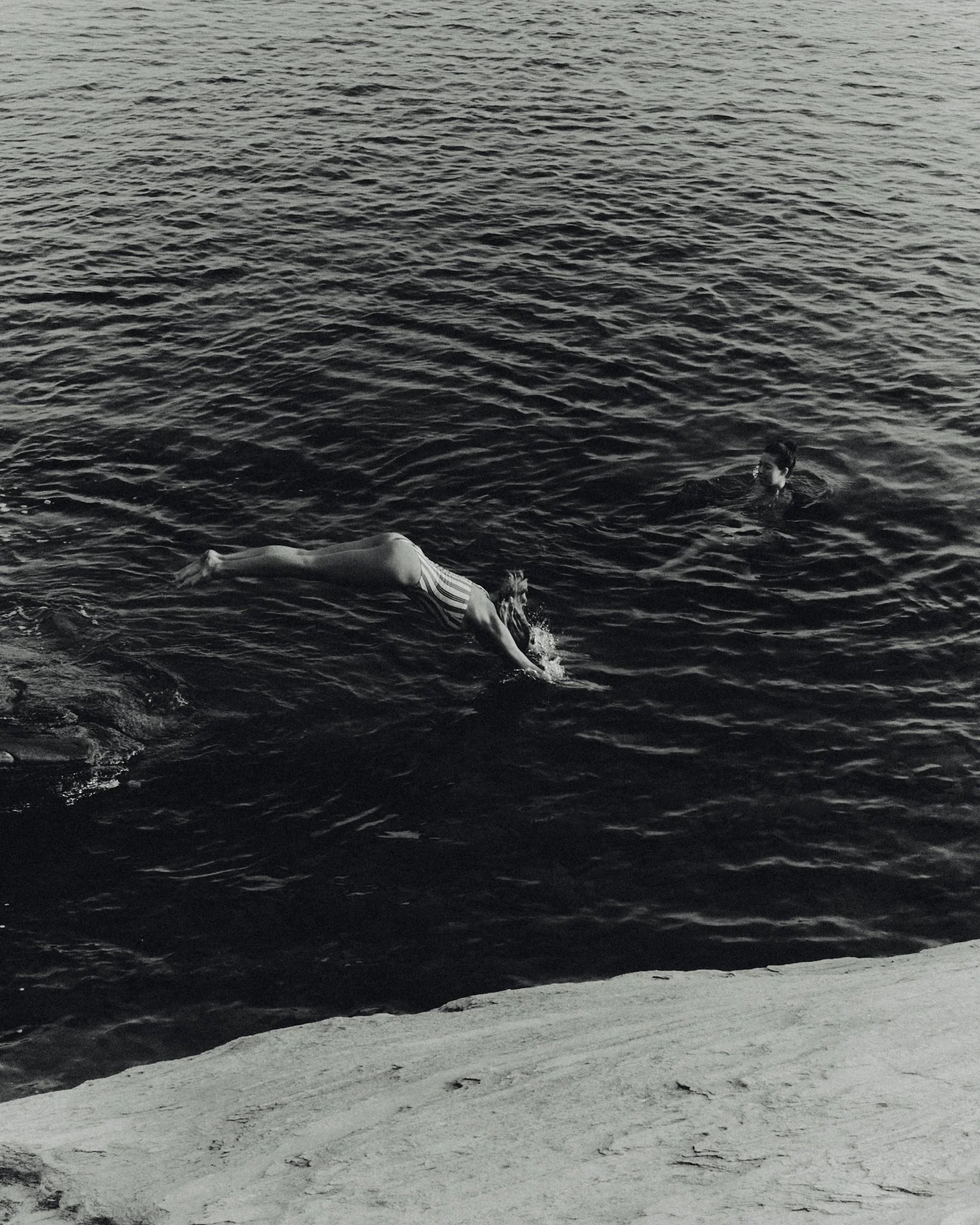 A person in a striped swimsuit swimming in a body of water near a rocky shore, with another person swimming nearby.