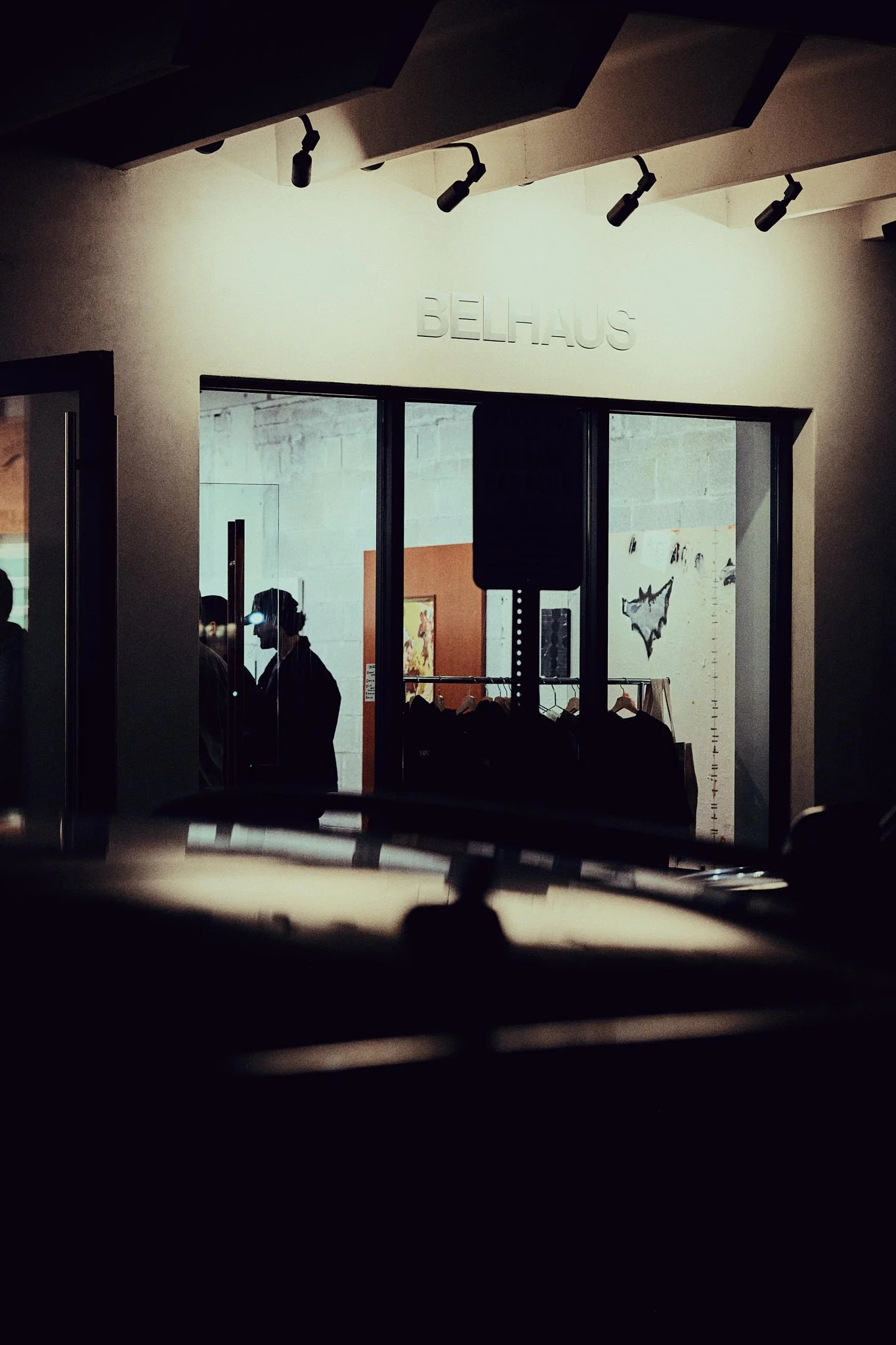 View through a car windshield showing an interior space called Belhaus with people talking, some wearing virtual reality headsets, and clothing racks inside.