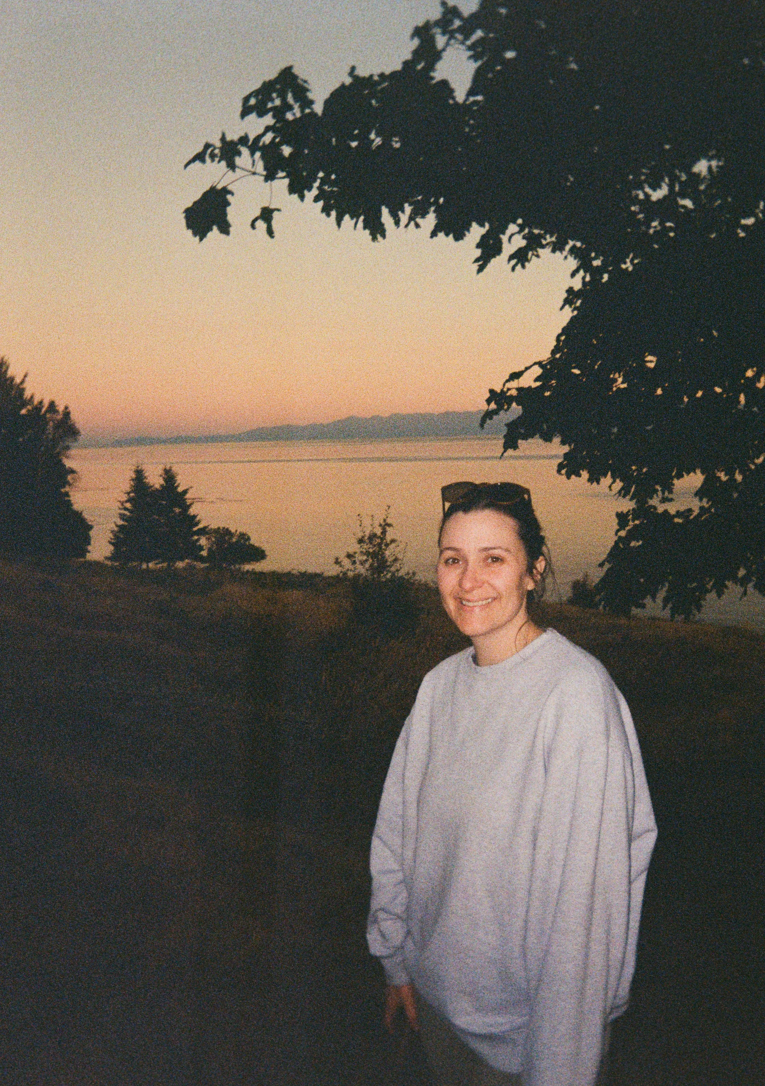 A woman smiling outdoors at sunset, standing near trees with a body of water in the background.