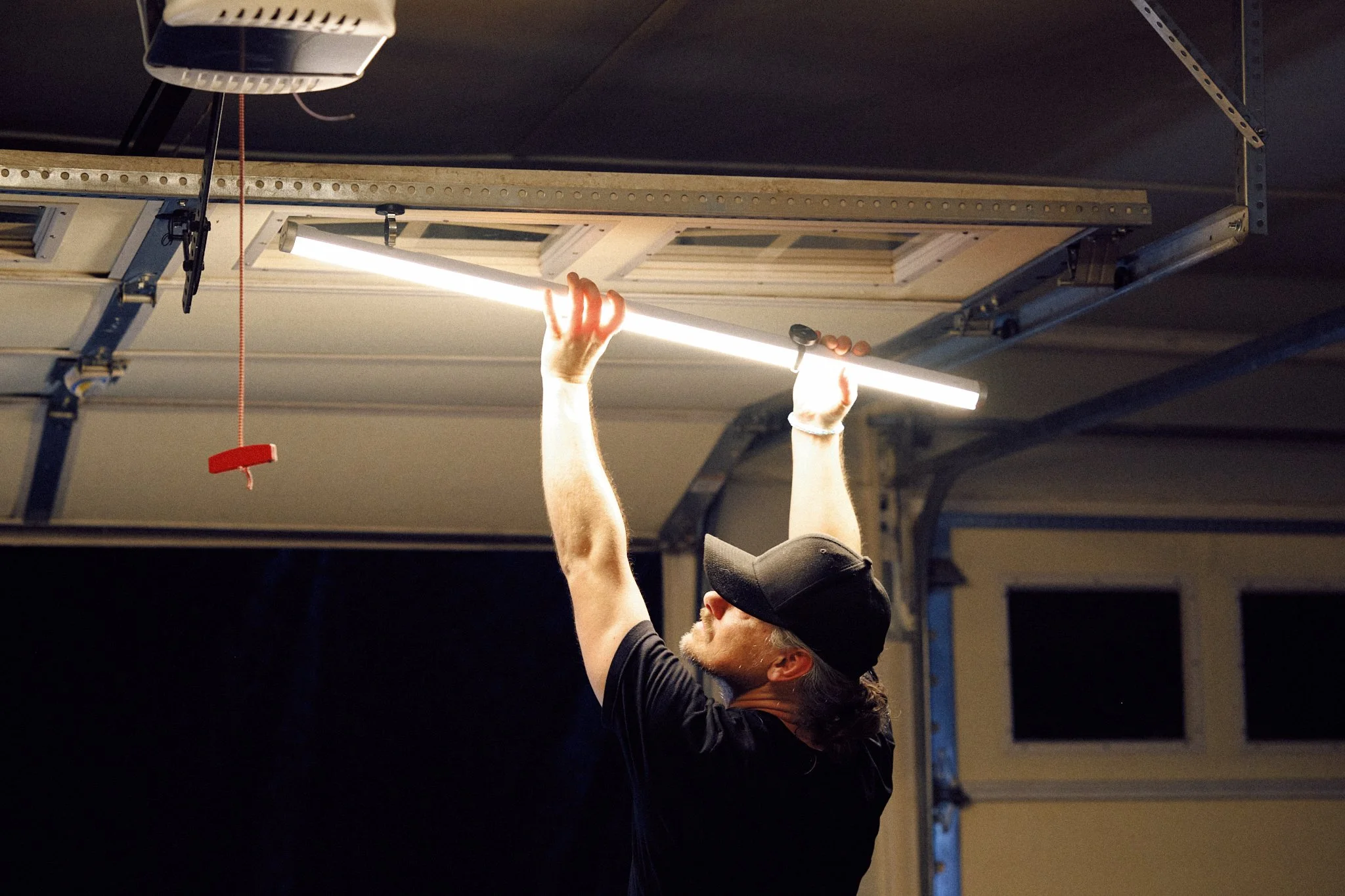 A man wearing a black cap and black T-shirt is installing or adjusting a fluorescent light fixture on a garage ceiling.