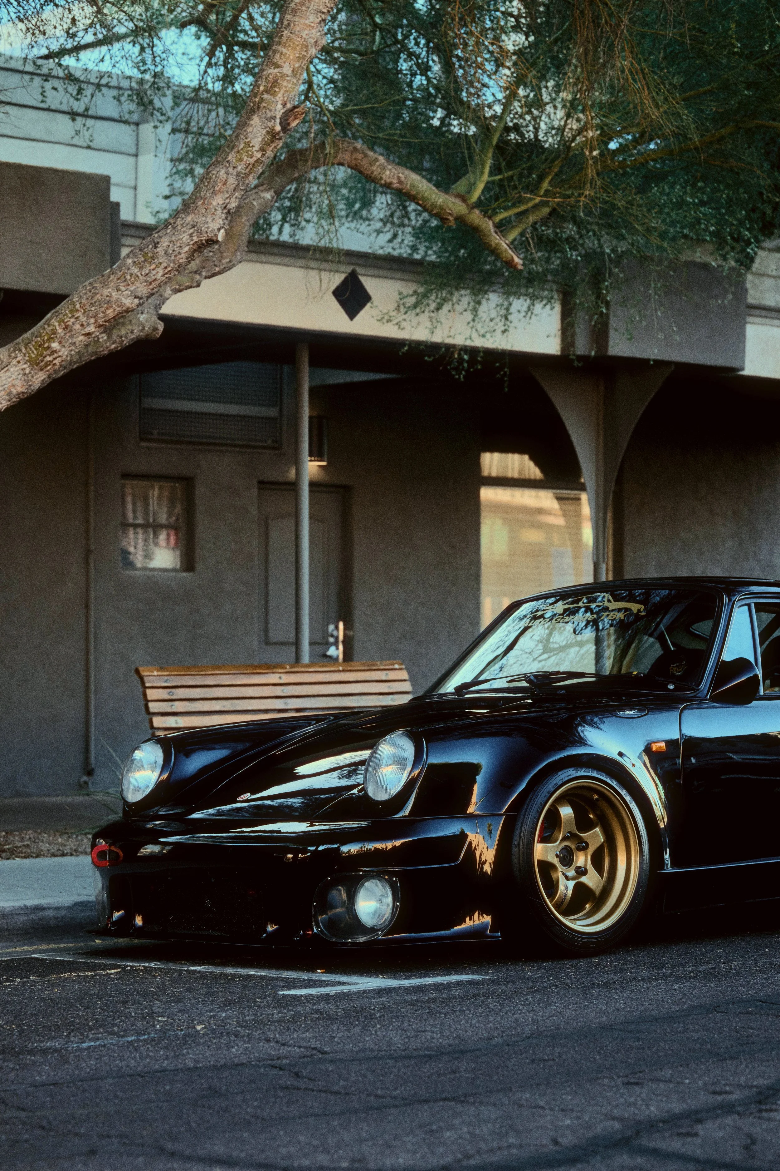 Black sports car with gold wheels parked on the street near a building with a bench and trees in the background.