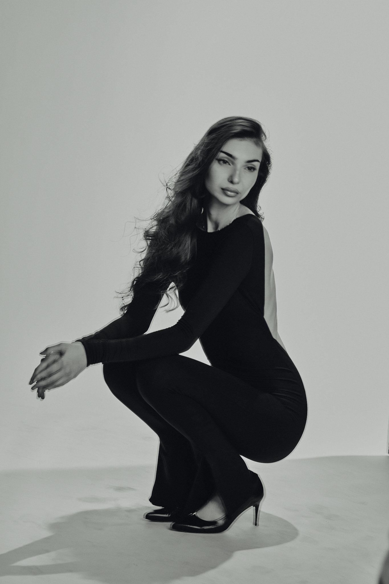 Black and white photo of a woman squatting in high heels, wearing a form-fitting long-sleeve dress with open back, gazing thoughtfully to the side.