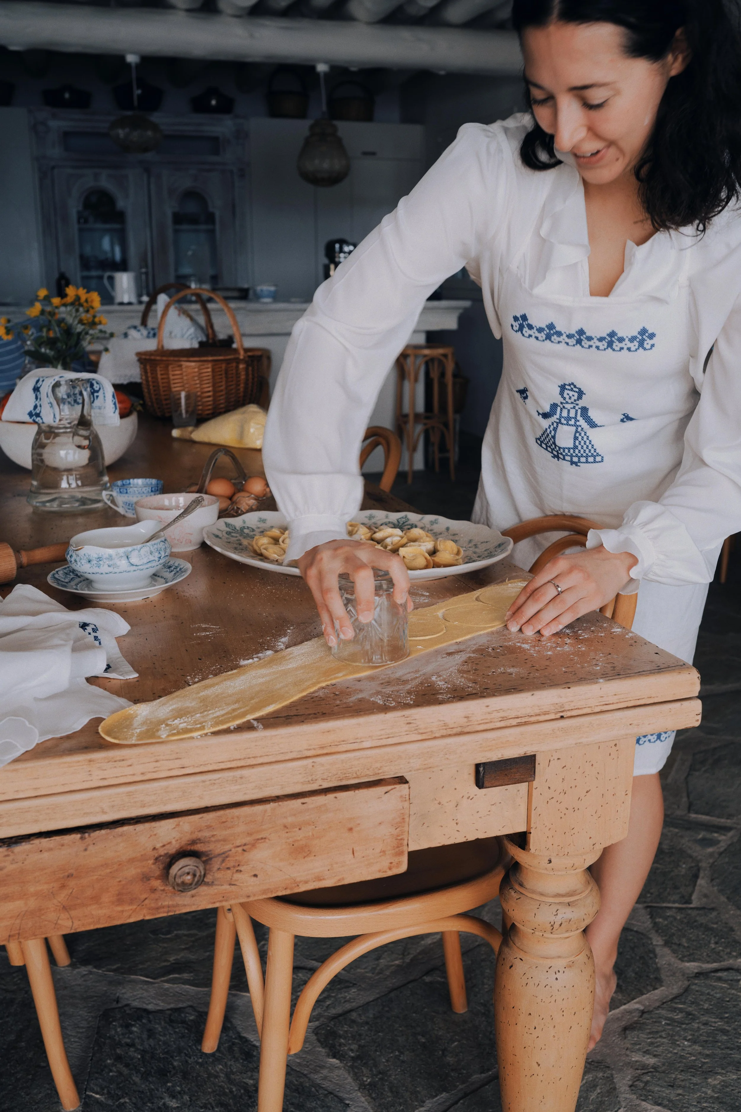 Woman in white dress with embroidered blue figure, rolling out pasta dough on a wooden table in a rustic kitchen.