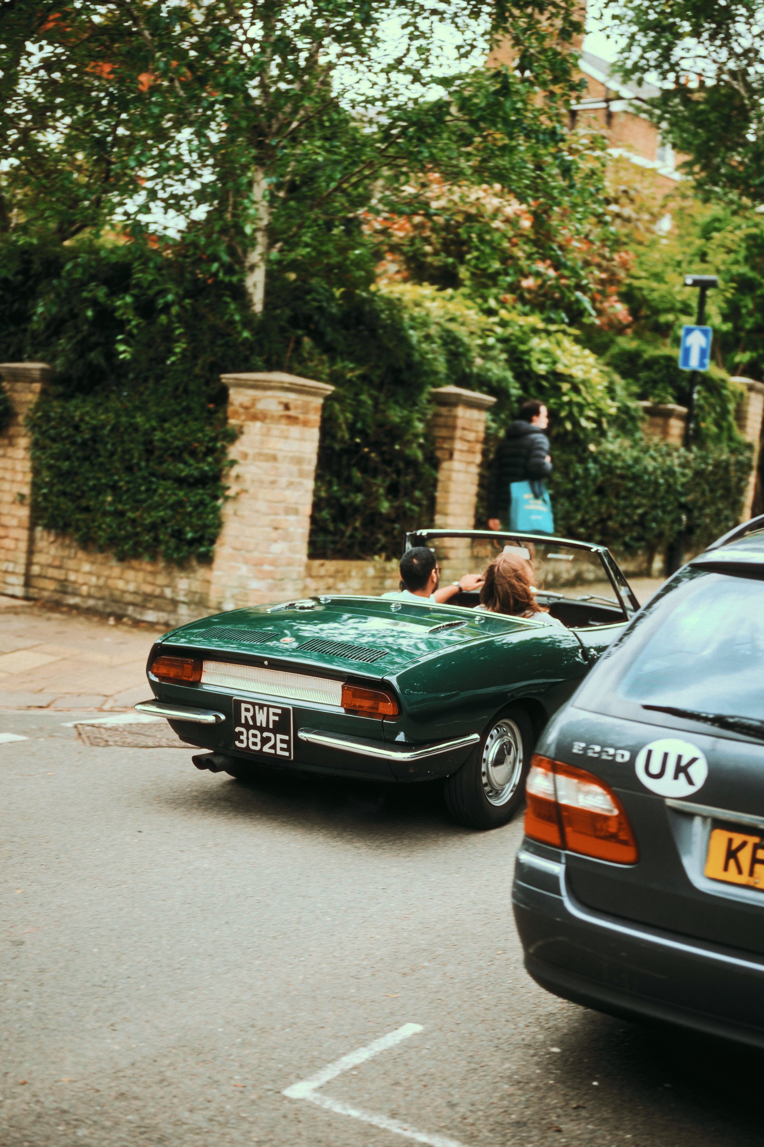A classic green convertible car with a license plate reading RWF 382E parked on the street. Two people are sitting inside the car, and a pedestrian is walking on the sidewalk nearby. The background features trees, a brick wall, and a blue one-way str