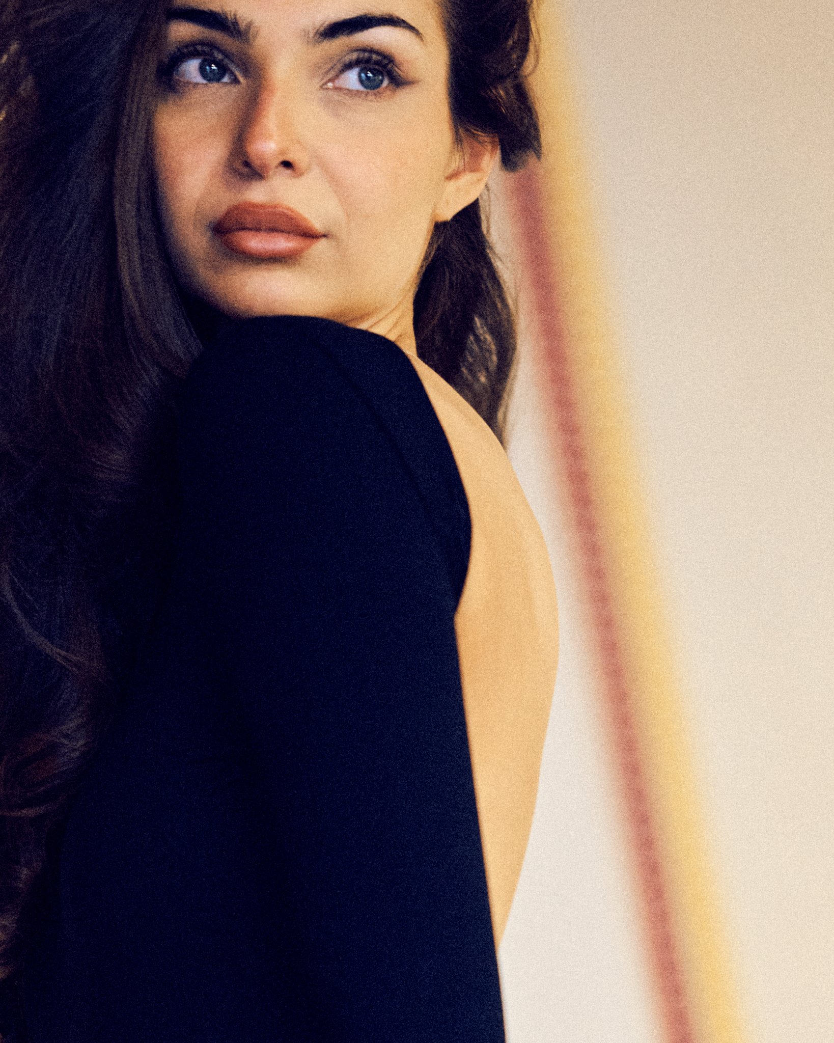 Close-up of a woman with dark hair and blue eyes, looking over her shoulder, wearing a black top.