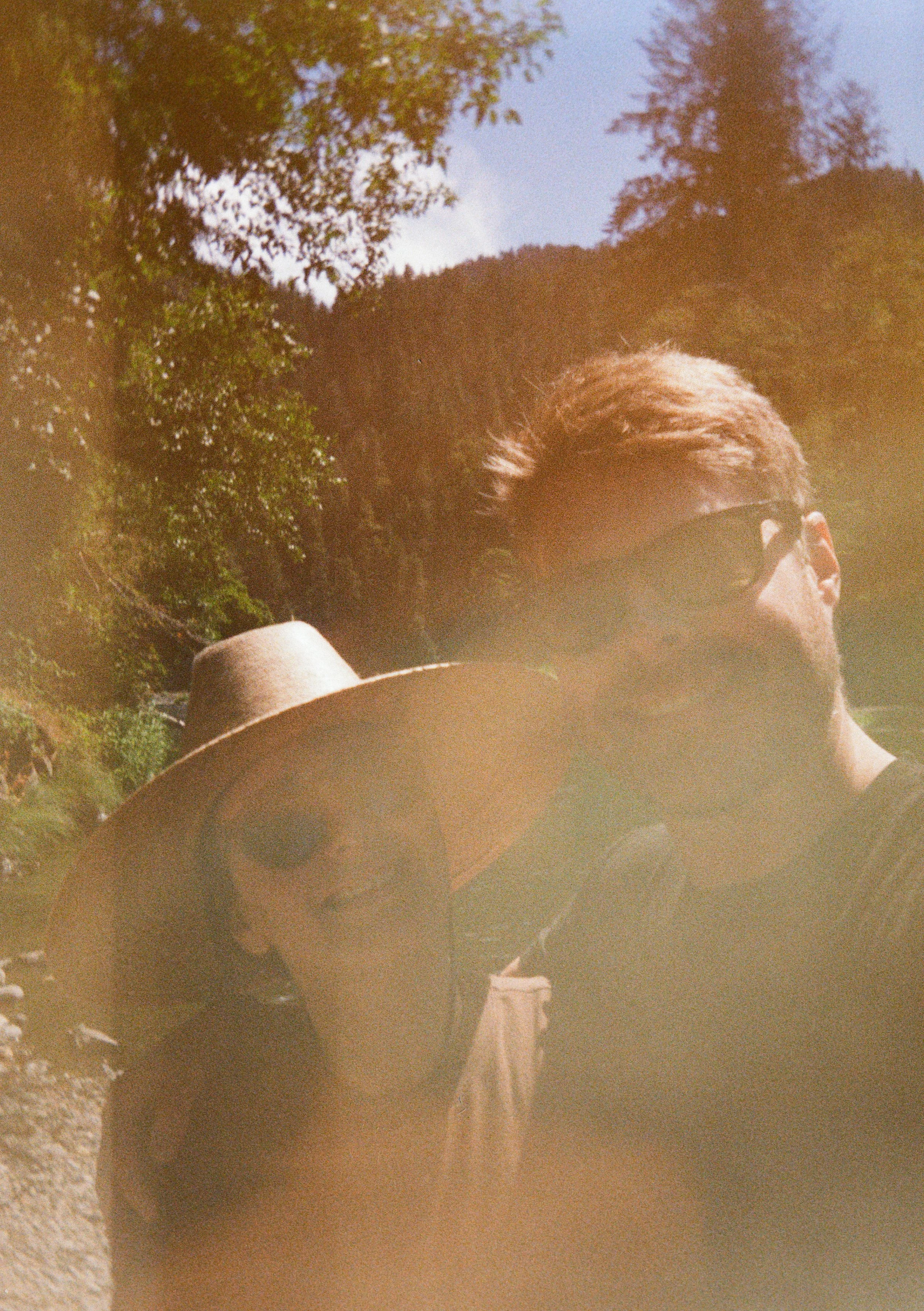 A man and a woman outdoors in a lush, mountainous area, both wearing sunglasses, with the man taking the selfie.