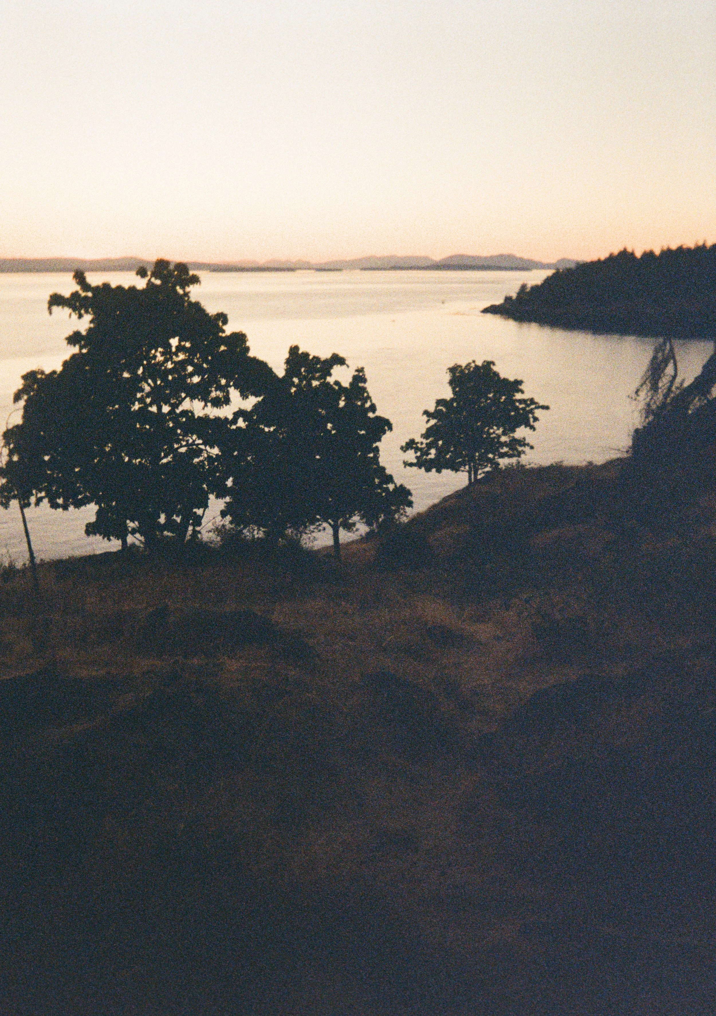 A serene lakeside view during sunset with trees lining the shore and with a distant mountain range.