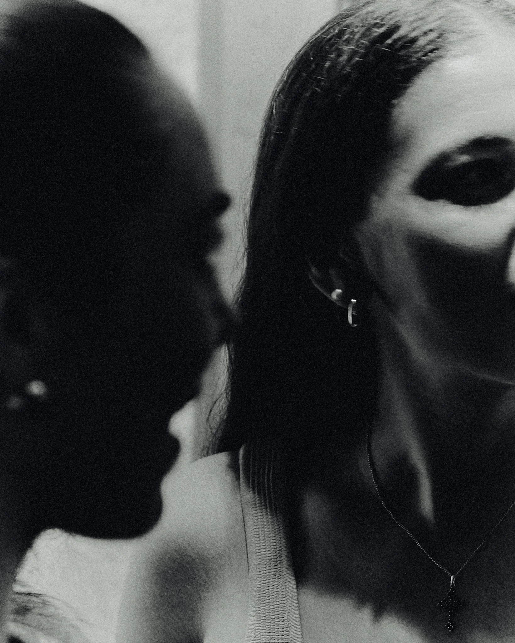 Close-up black and white photo of two women, one wearing a mask, both with earrings and necklaces, with one woman in focus and the other blurred.