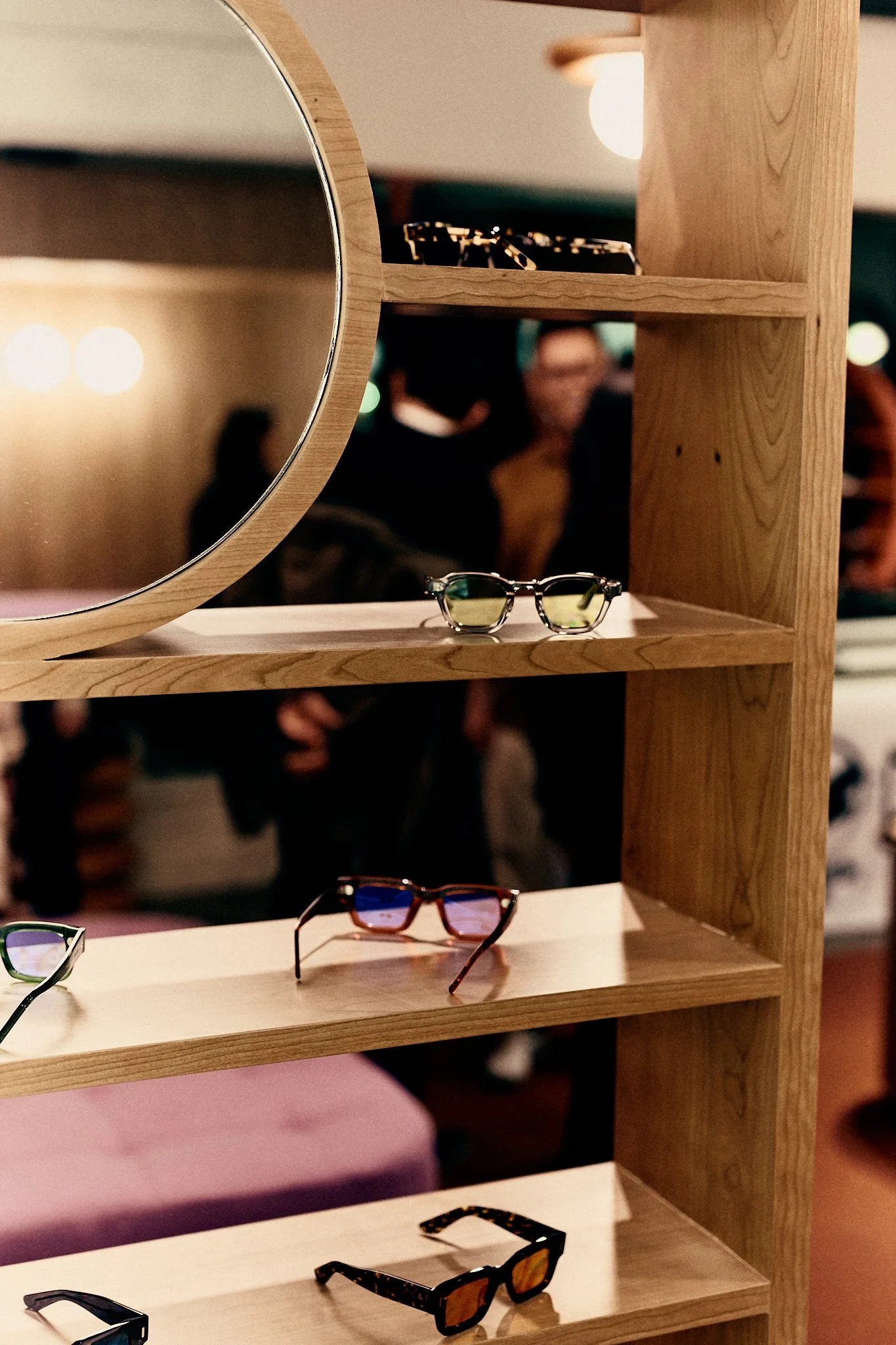 A wooden display shelf with various sunglasses on each tier, seen through a partial mirror. Blurred background of people at an indoor event.