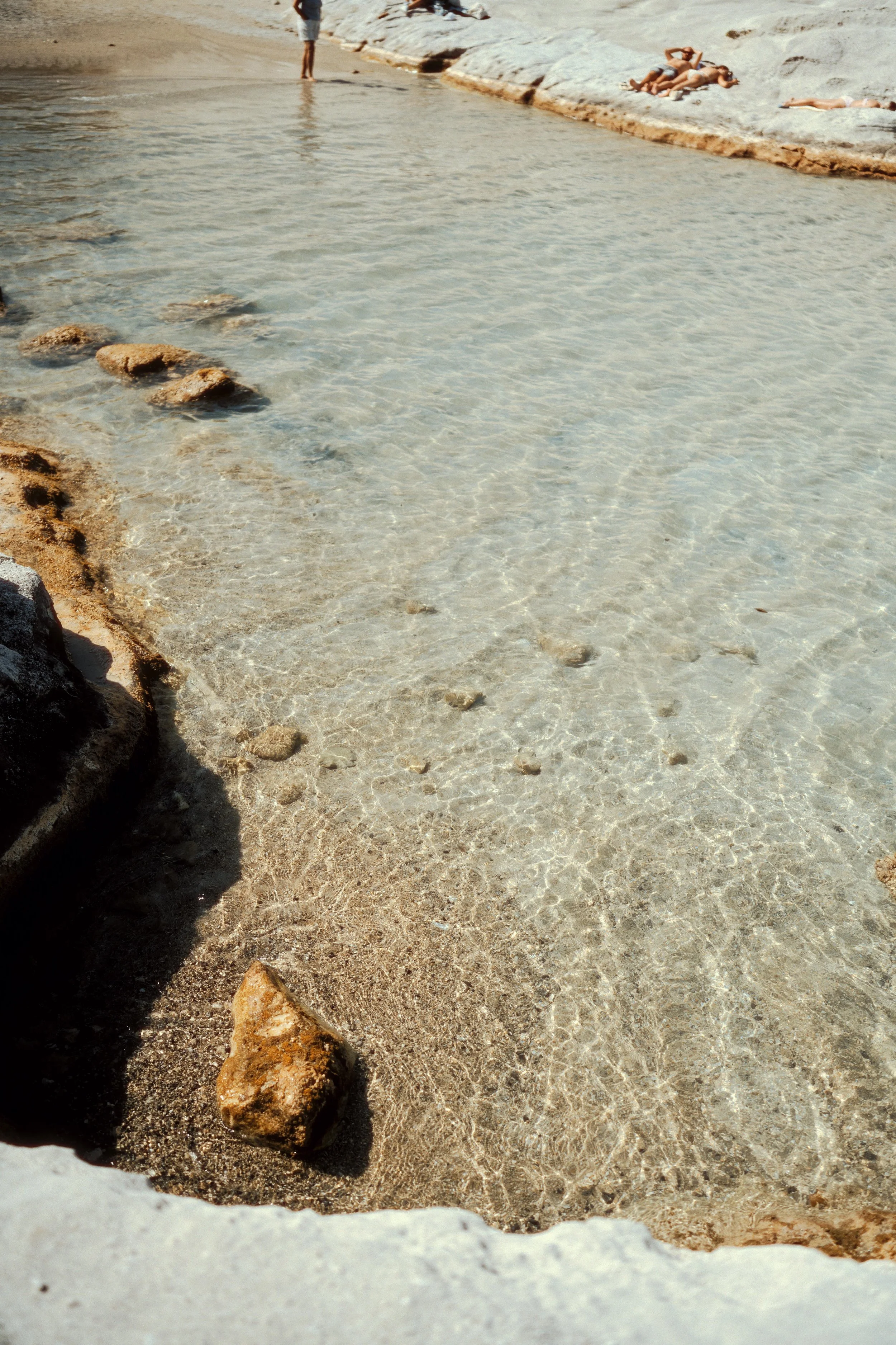 Clear, shallow water with rocks and sand, people sunbathing on white rocks, and some individuals standing at the water's edge.