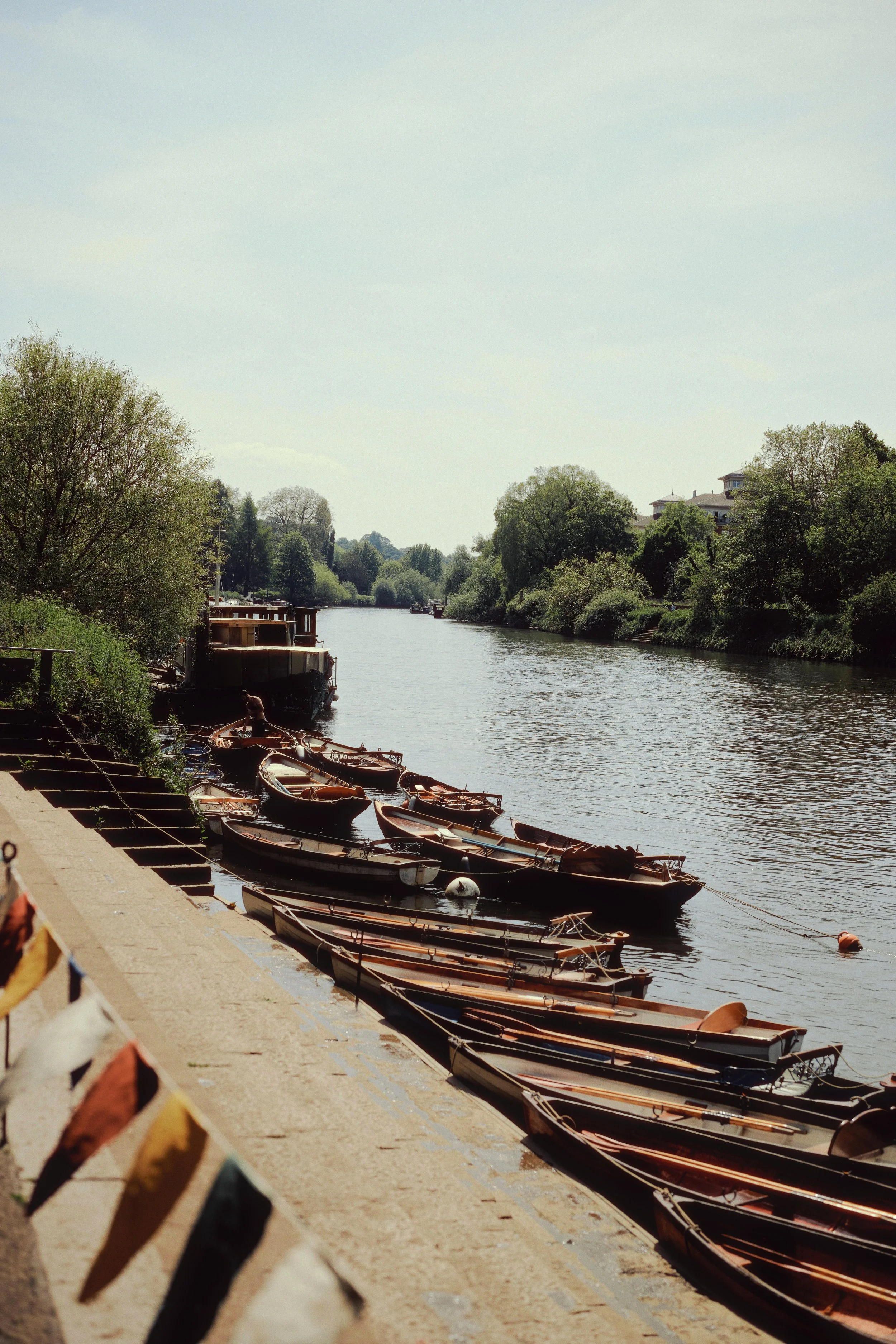 Boats moored along a riverbank on a sunny day with trees and houses in the background.