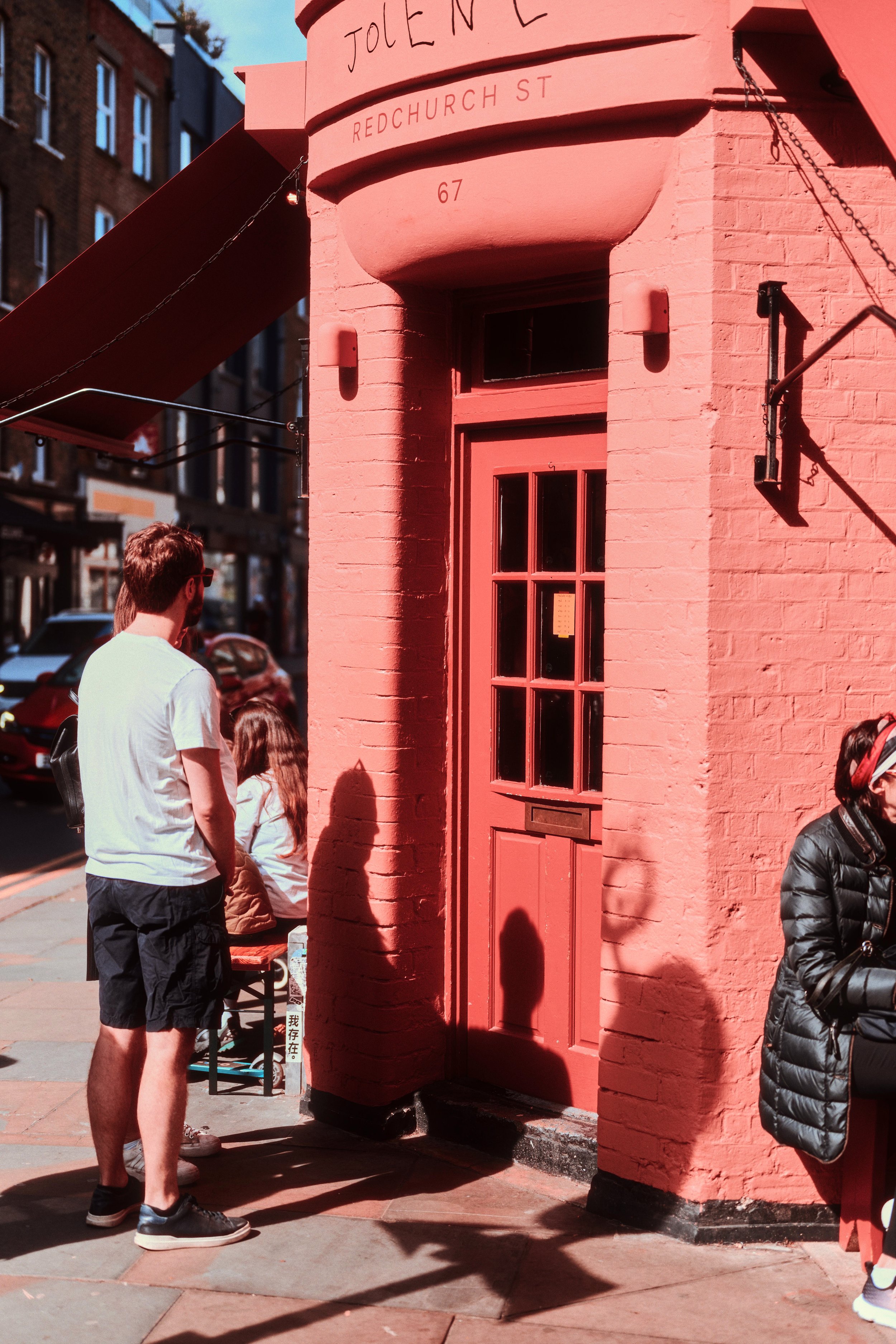 A street corner with a pink brick building labeled 'JOLENE' and 'REDCUHRCH ST 67,' with people standing outside on the sidewalk.