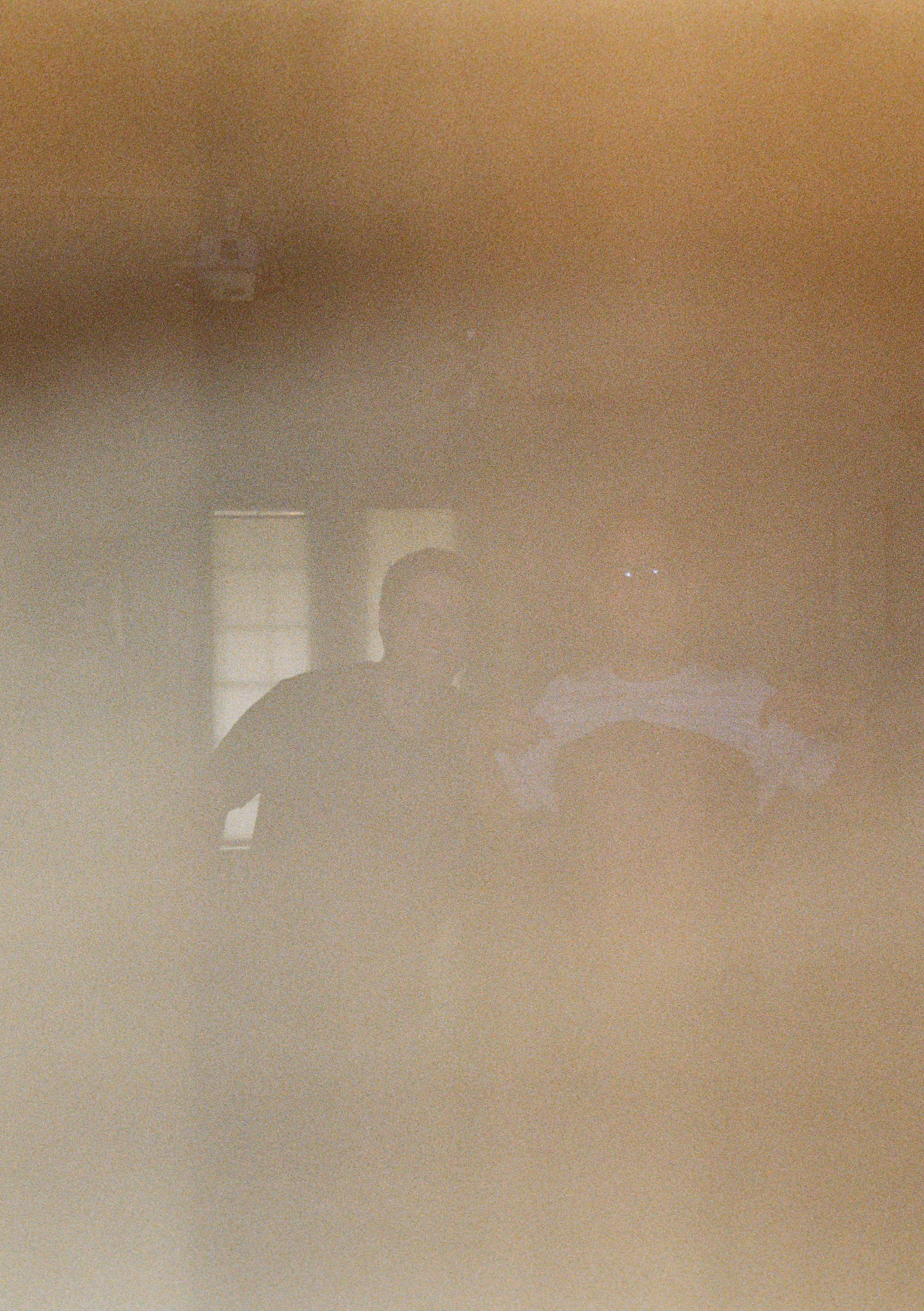 A foggy or smoky scene with two people sitting at a table, engaged in conversation in an indoor environment.