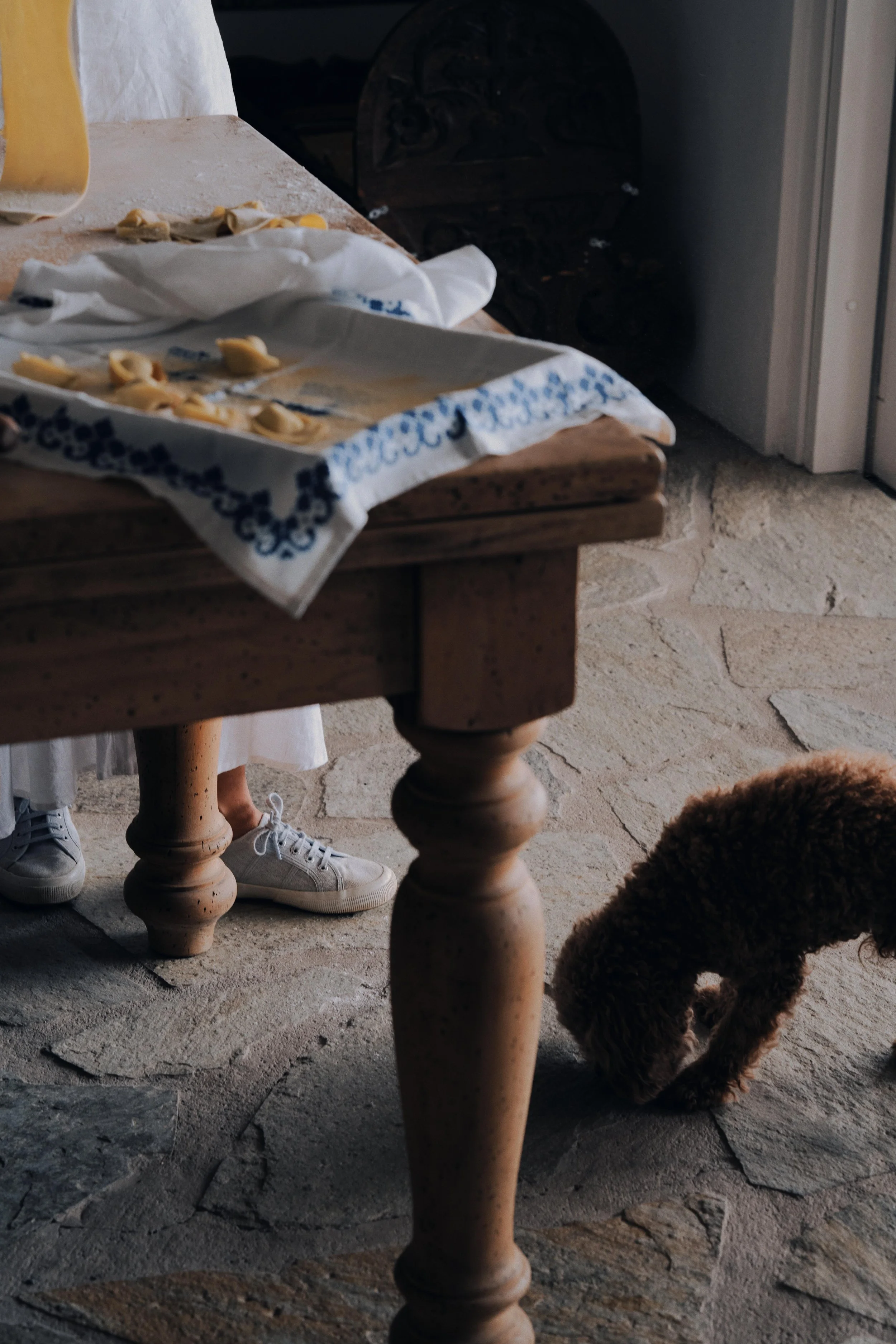 A rustic kitchen scene with a wooden table and stone floor. There are unfinished fresh pasta shapes on a cloth. Part of a person's legs and white shoes are visible behind the table, and a small curly-haired brown dog is sniffing the floor nearby.